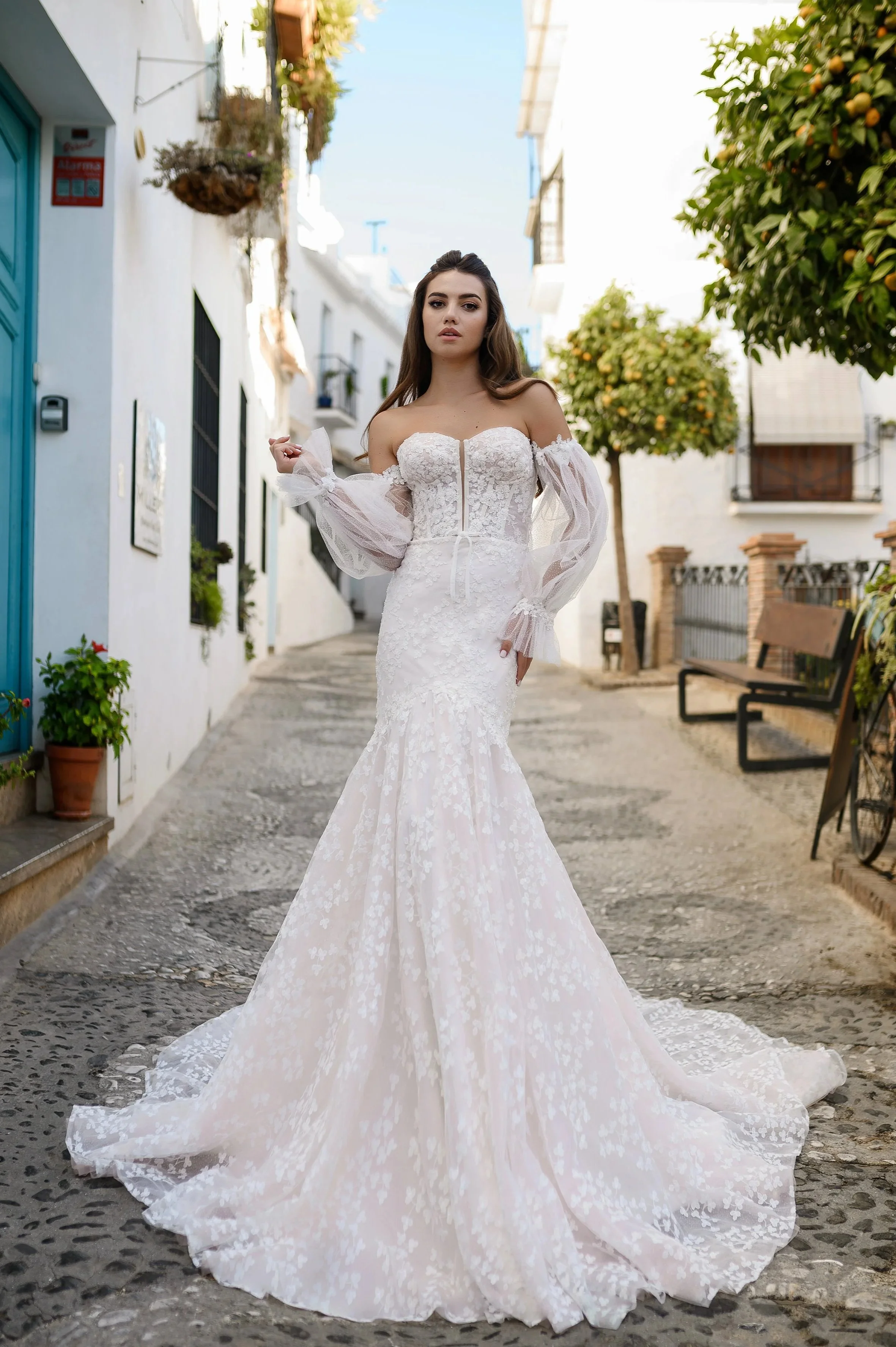 Une femme en robe de mariage blanche dans une rue pavée, entourée de maisons blanches avec des fenêtres ornées de plantes et d'arbres fruitiers.
