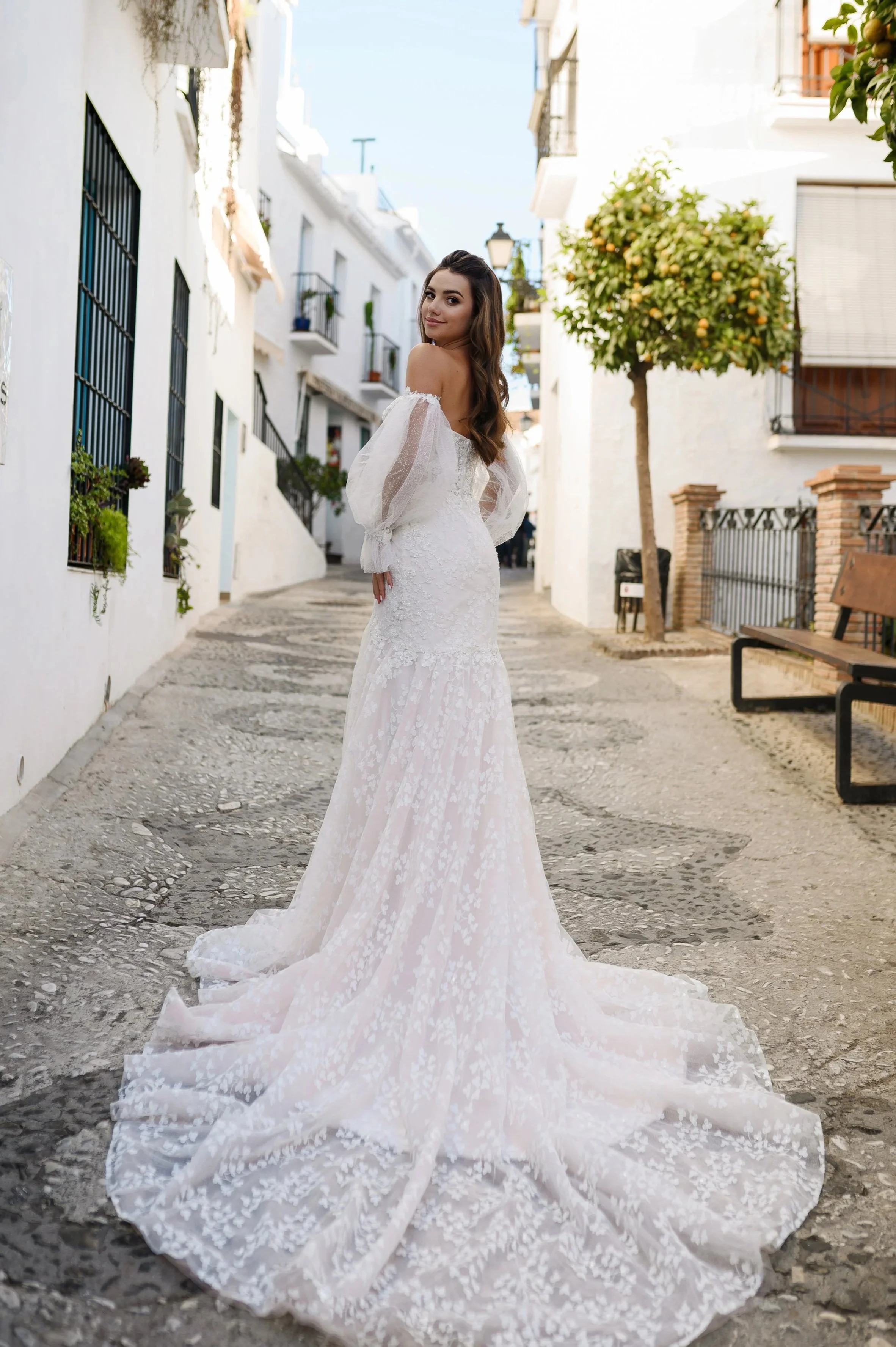 Une femme en robe de mariée blanche pose dans une ruelle pavée à cheval entre une vieille ville ensoleillée et ses bâtiments blancs.