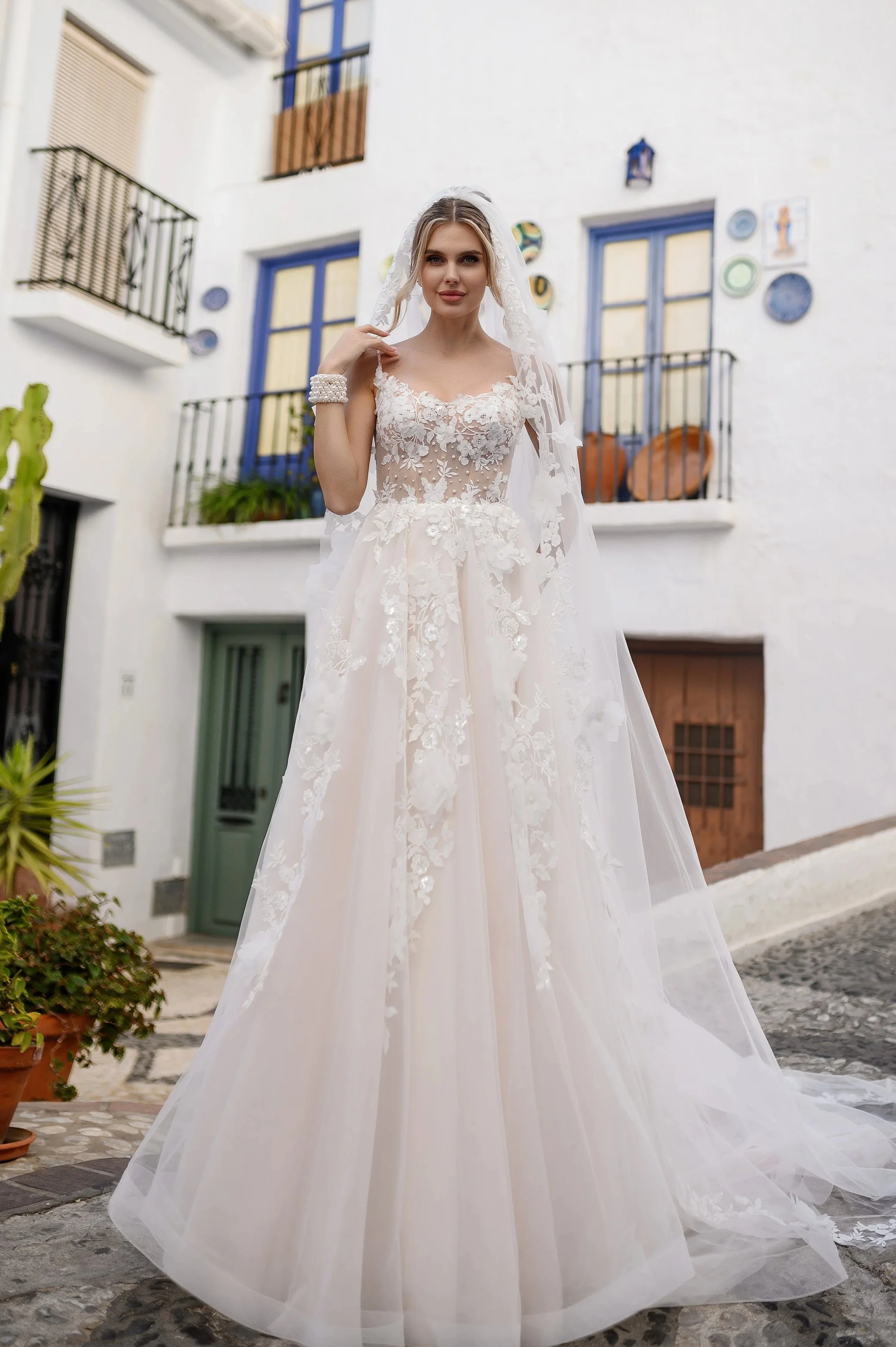 Femme en robe de mariage blanche avec dentelle, à l'extérieur devant une maison blanche avec balcons et décoration colorée.