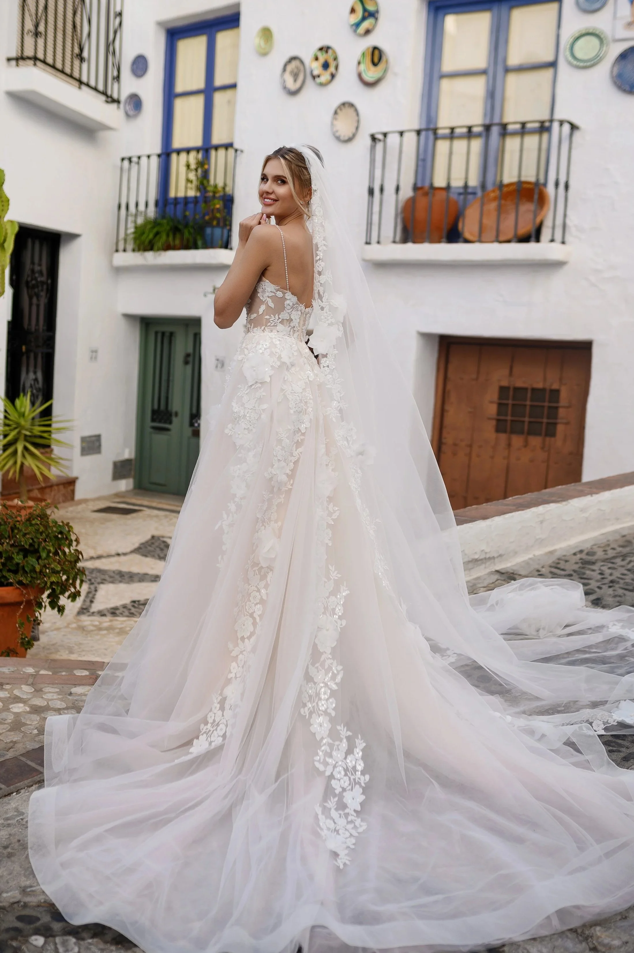 Une jeune femme en robe de mariée blanche avec des broderies florales, se tient dans une rue pavée devant un bâtiment blanc avec des volets bleus et des plantes en pot.