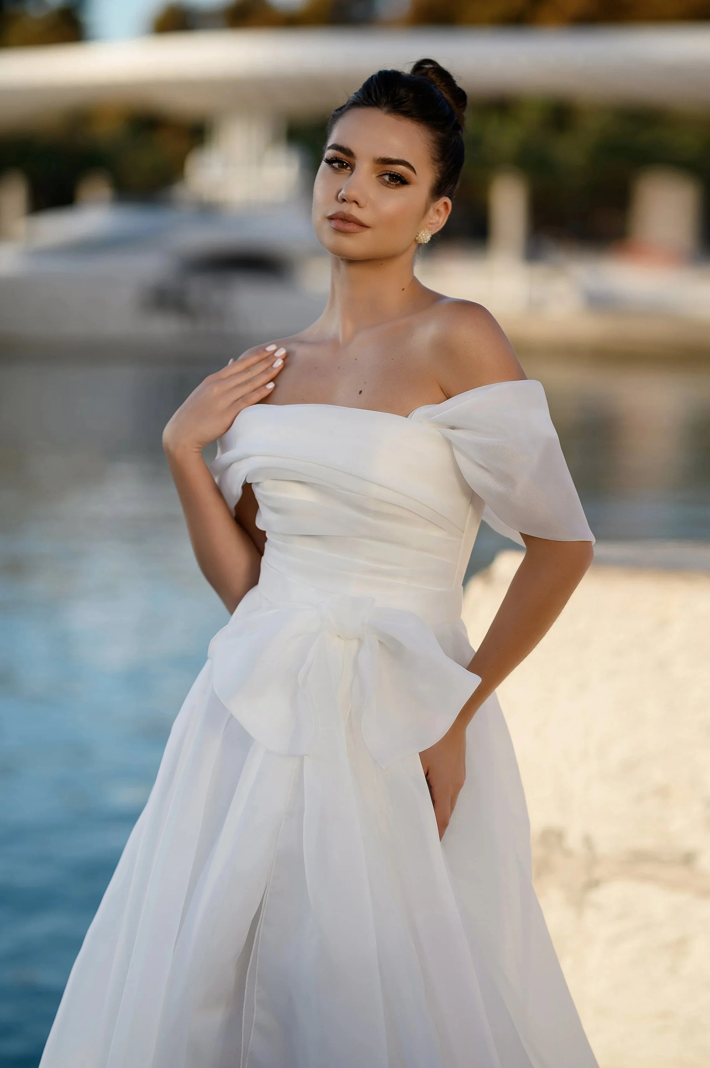 Une femme en robe de mariée blanche pose près d'un plan d'eau avec un bateau en arrière-plan.