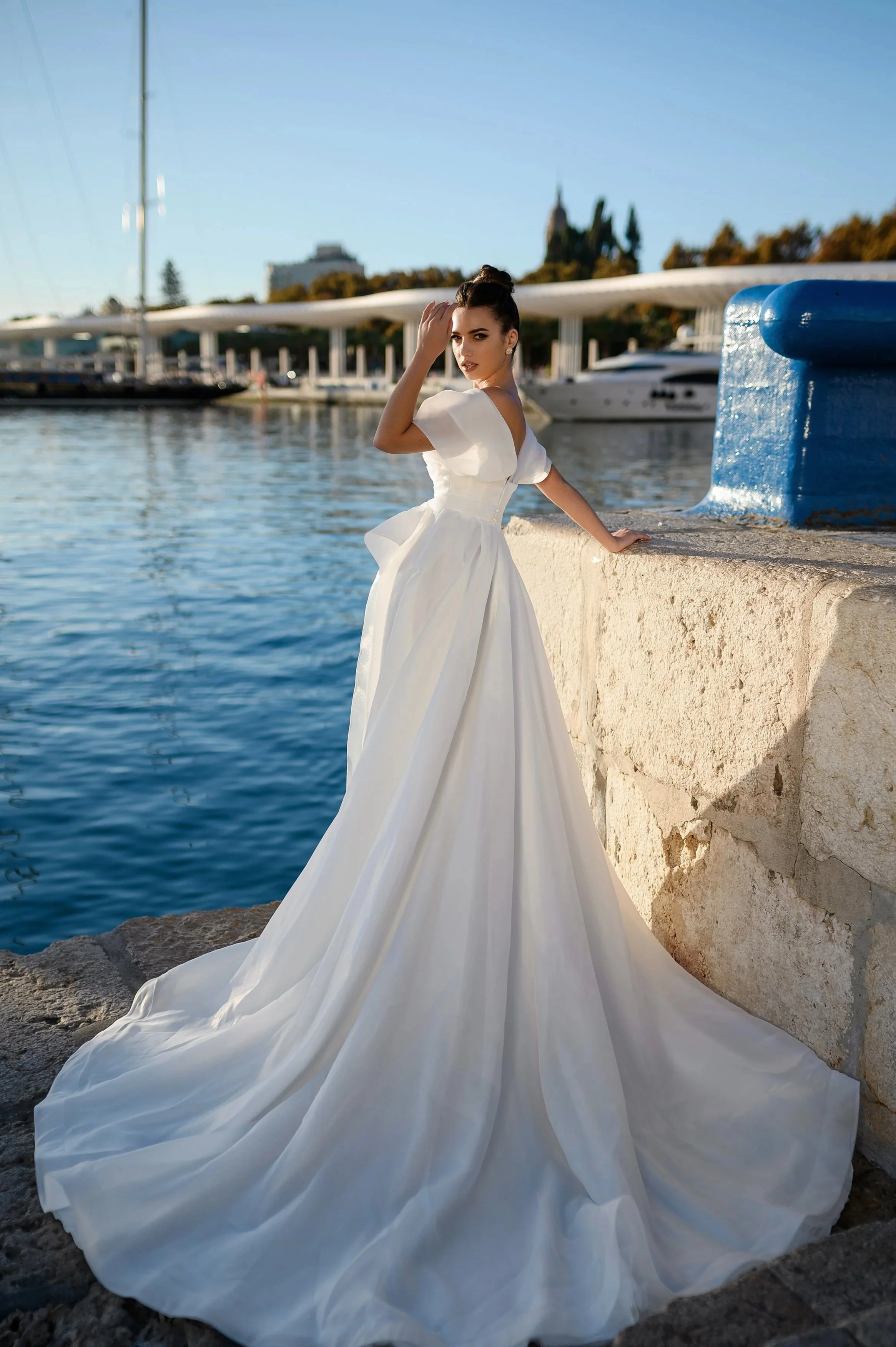 Une femme portant une robe de mariée blanche devant un port avec des yachts et un ciel clair.