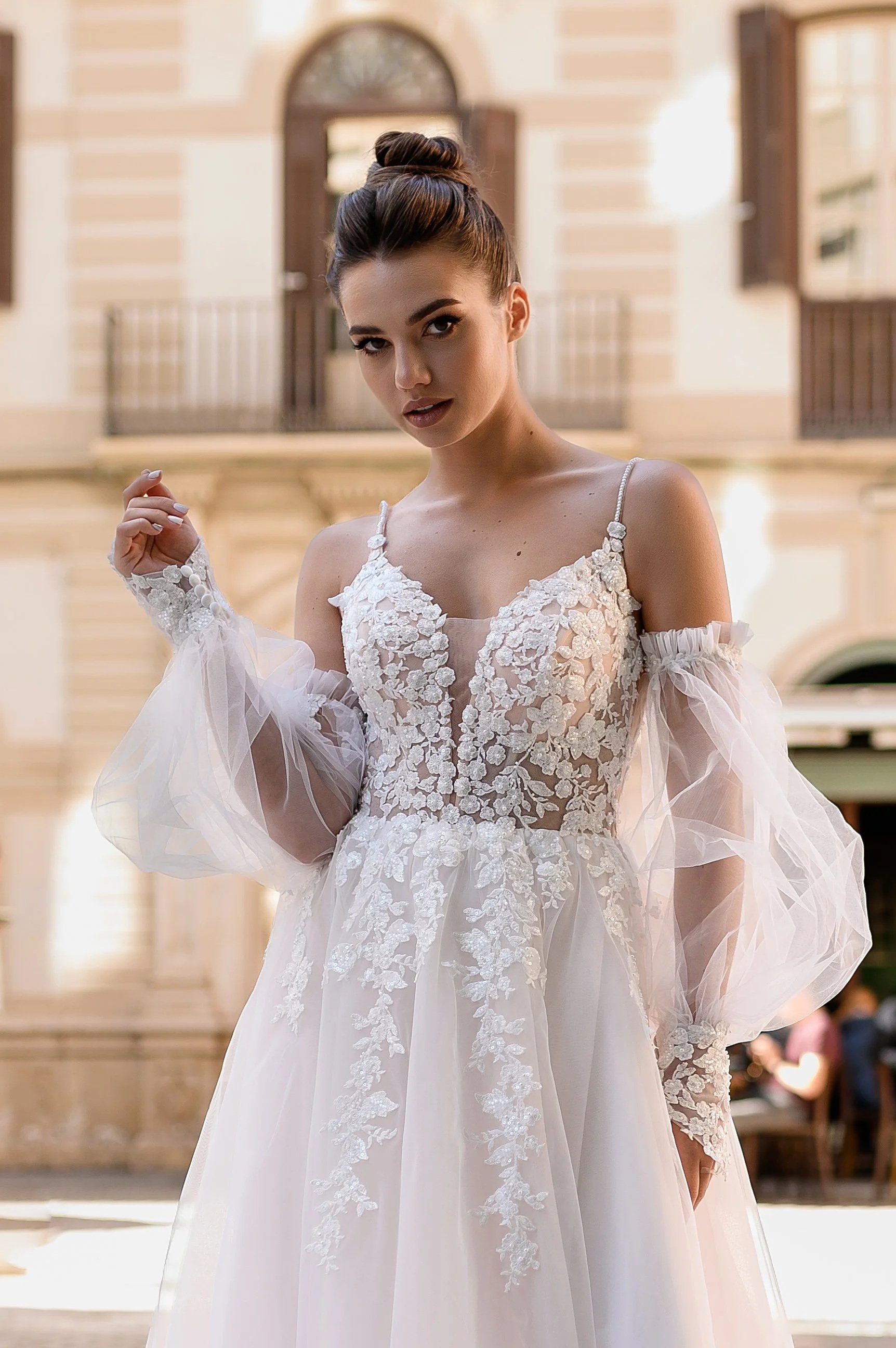 Une femme porte une robe de mariée blanche avec des détails en broderie florale, devant un bâtiment en pierre avec des fenêtres et des balcons en bois.