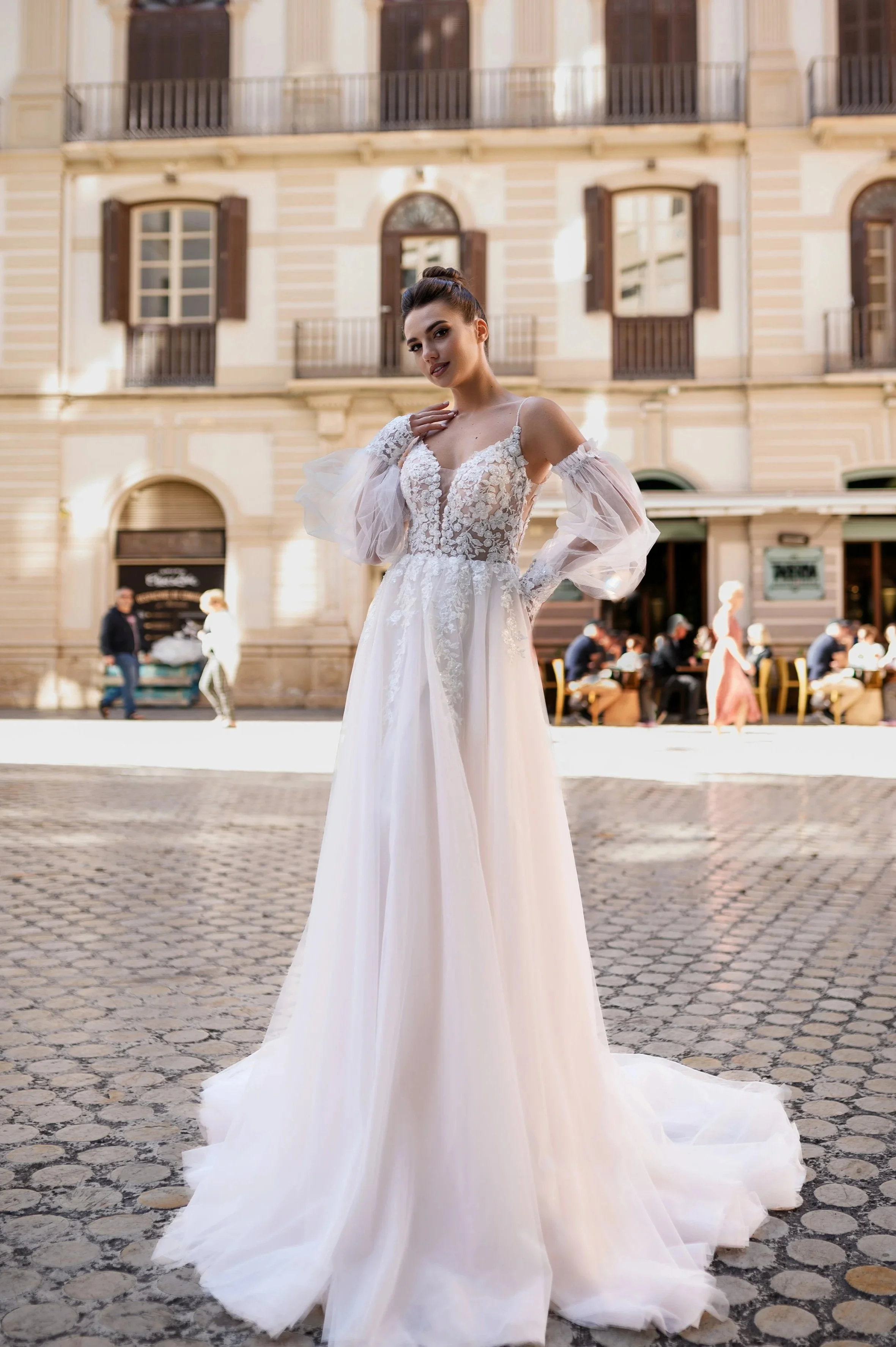 Une femme en robe de mariage se tenant dans une rue pavée en ville, avec un bâtiment ancien en arrière-plan, en plein jour.