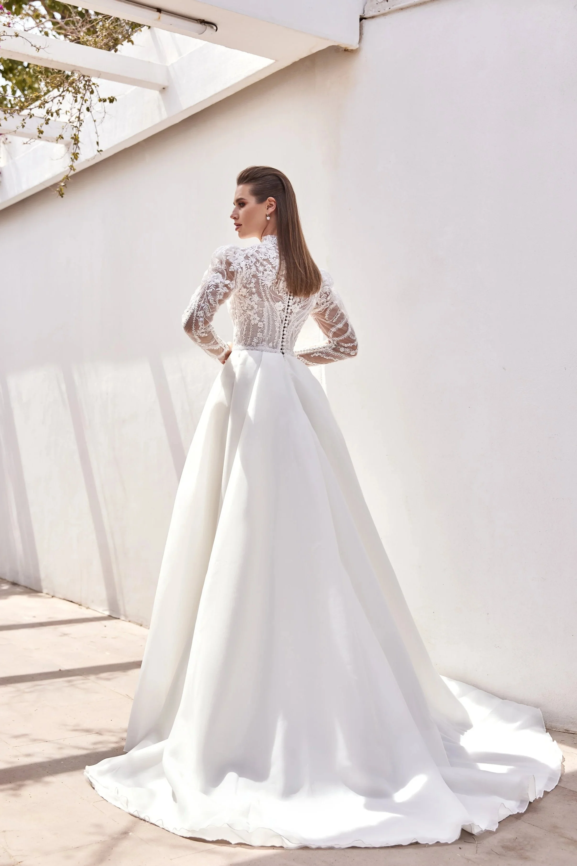 Une femme portant une robe de mariée blanche avec un haut en dentelle et une jupe en satin, debout contre un mur blanc dans une lumière naturelle.