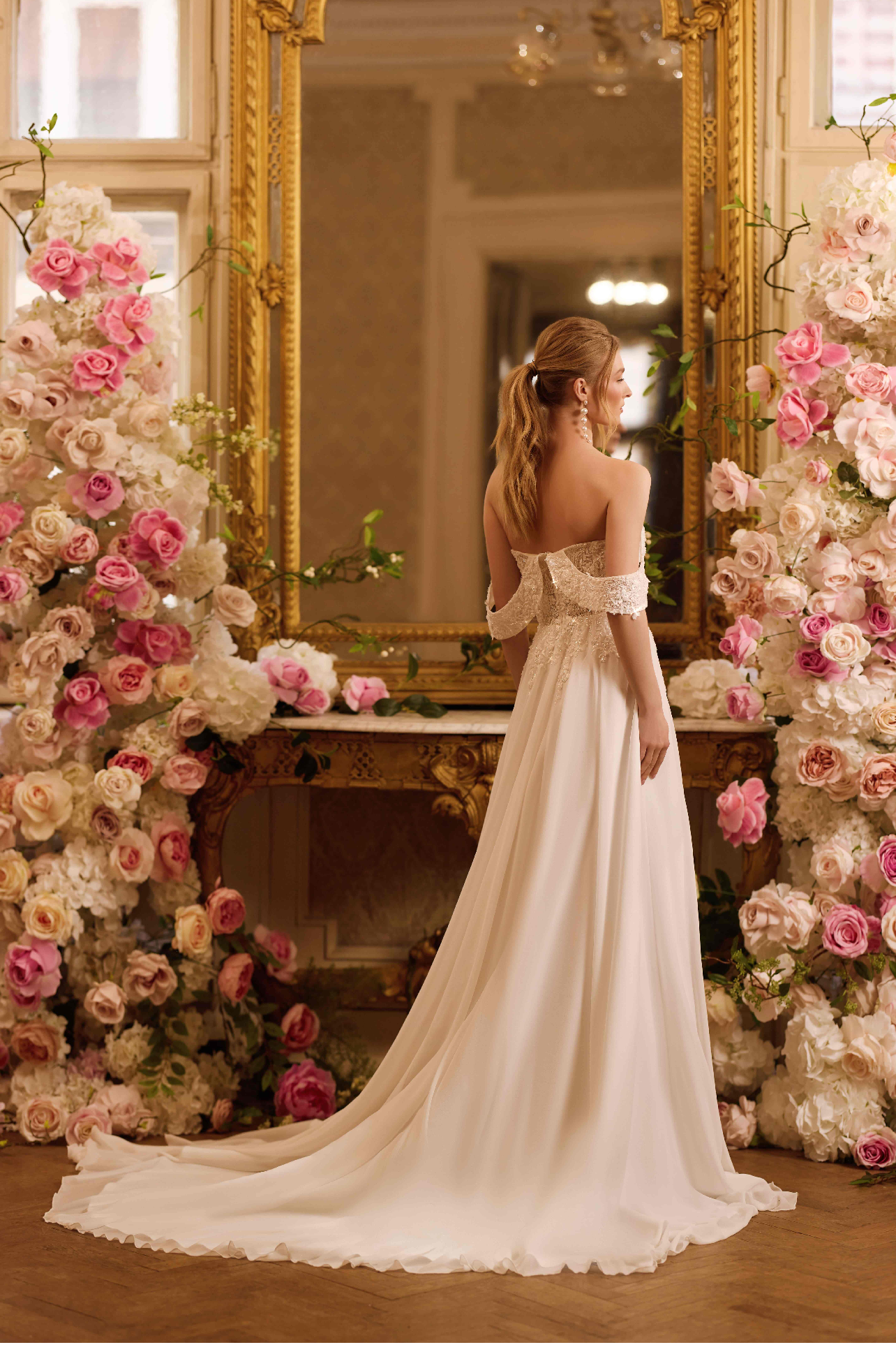 Une femme en robe de Mariée blanche élégante avec des épaules dénudées, se tient devant un grand miroir doré entouré de fleurs roses et blanches dans un décor luxueux.