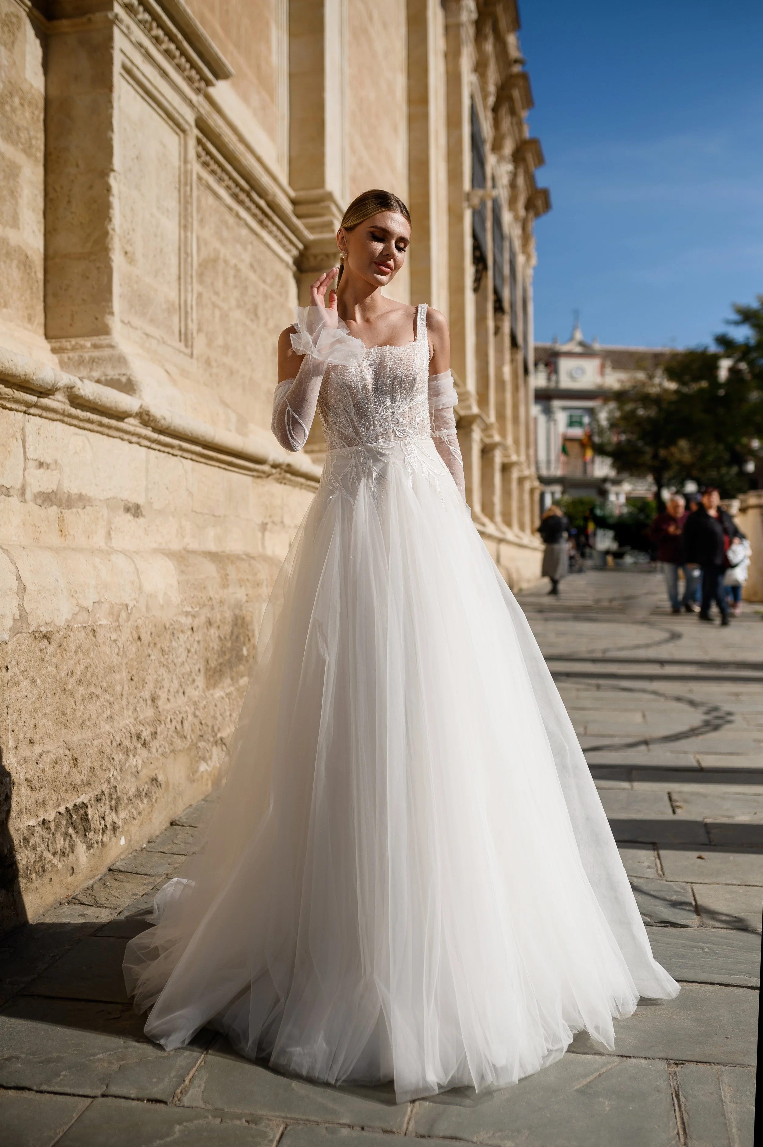 Une femme en robe de mariée blanche debout devant un mur en pierre, avec un ciel bleu en arrière-plan, dans un environnement urbain.