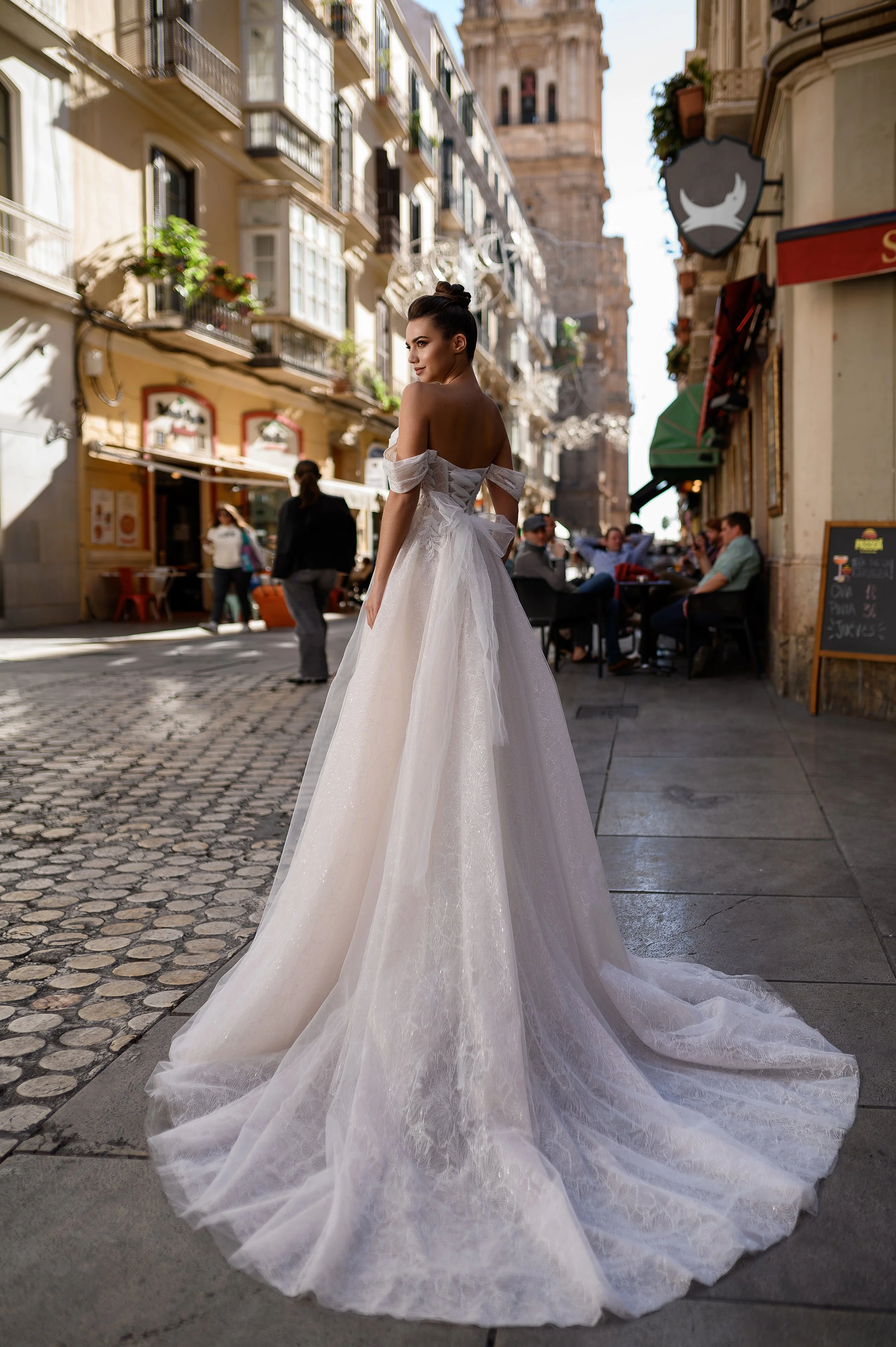 Femme portant une robe de mariée blanche dans une rue pavée, avec des bâtiments anciens et des cafés en arrière-plan.