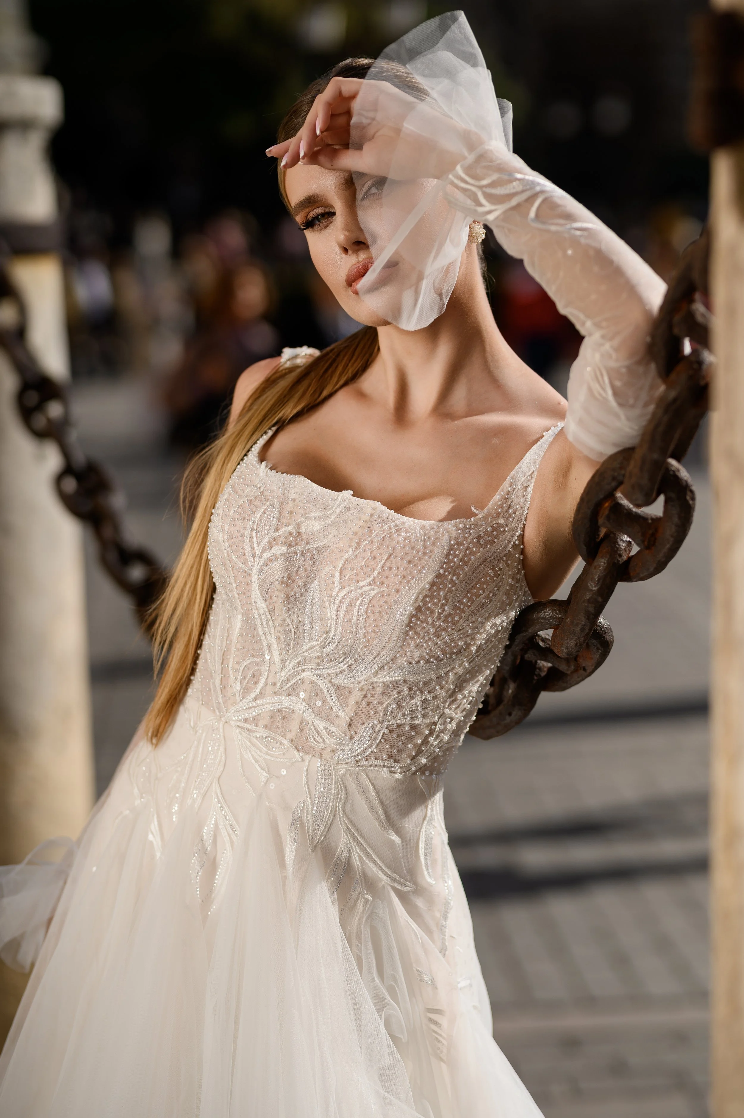 Une femme en robe de mariée blanche avec des détails en perles et broderies, portant un voile et des gants, posant près d'une chaîne en extérieur lors du coucher du soleil.