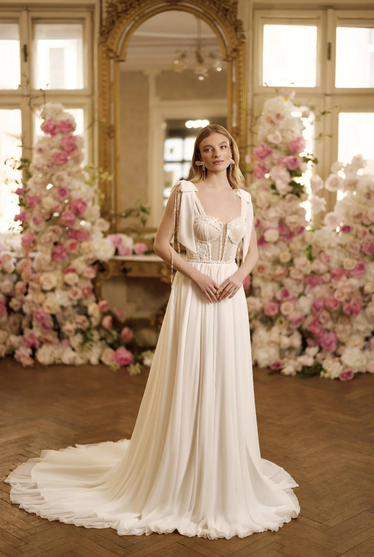 Une jeune femme portant une robe de mariée blanche dans une pièce décorée de nombreuses fleurs roses et blanches, avec un grand miroir ancien en arrière-plan.