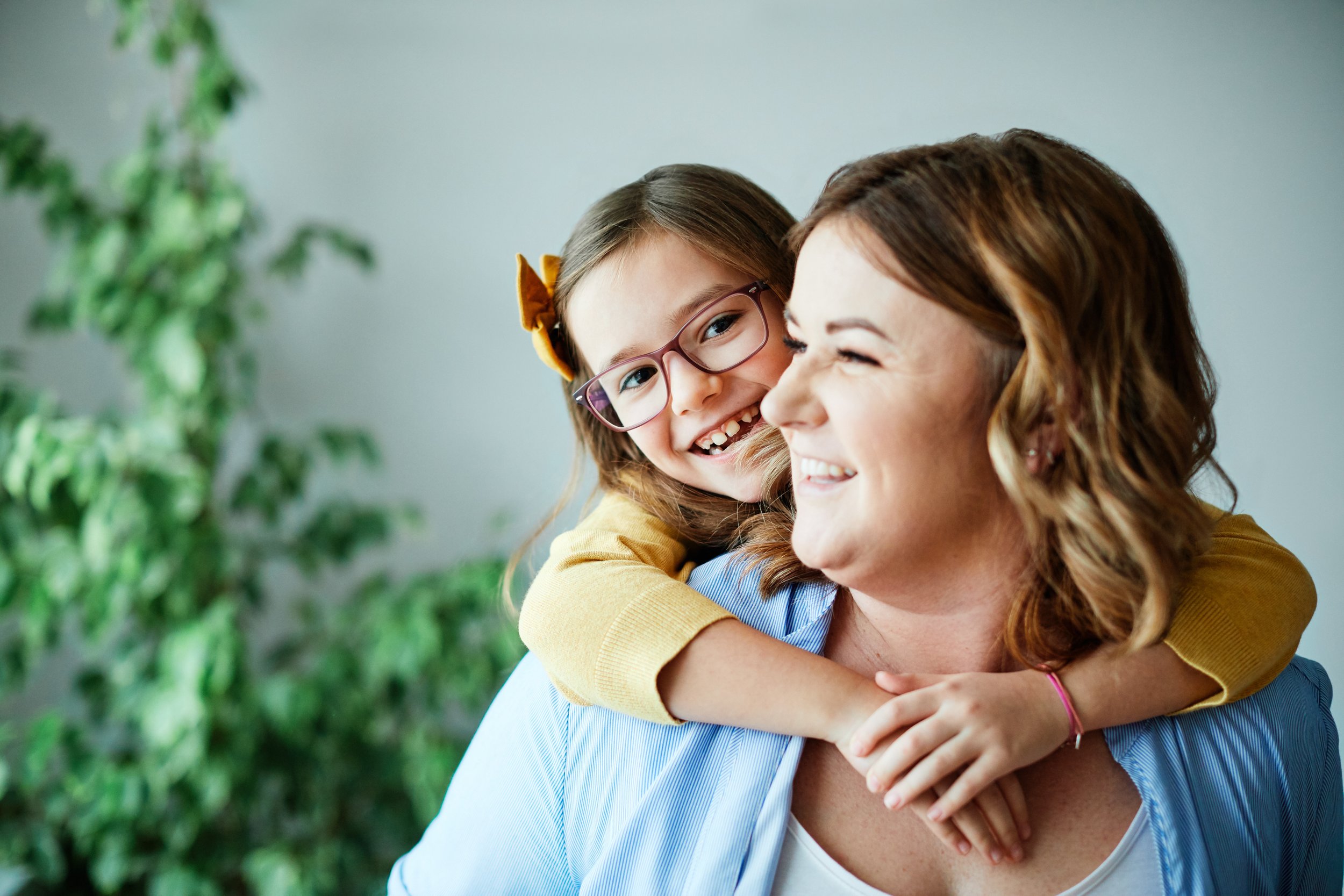 A mother gives her young daughter with glasses a piggy back ride as they both smile brightly.