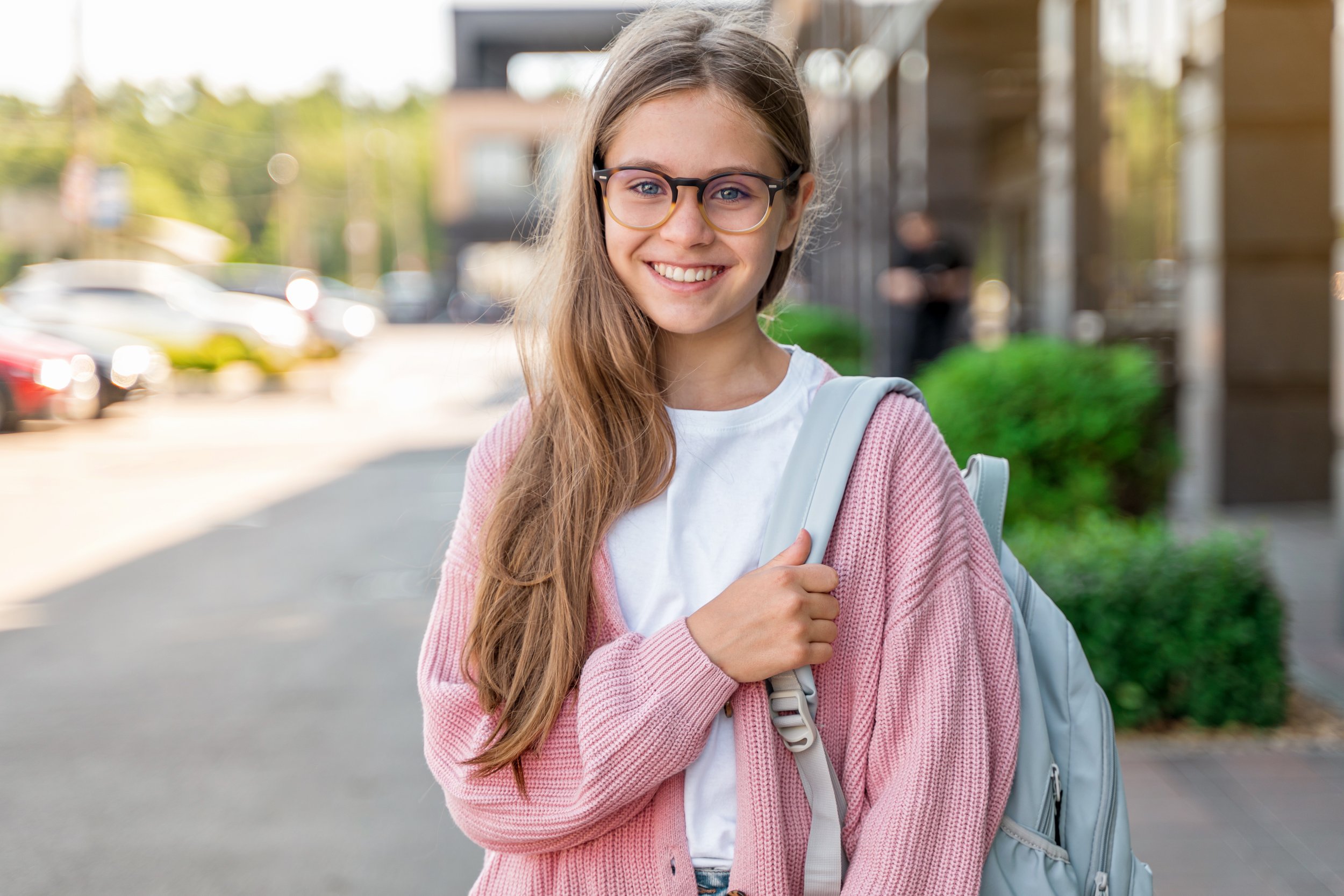 Teen girl in glasses and pink sweater standing outside school with a backpack