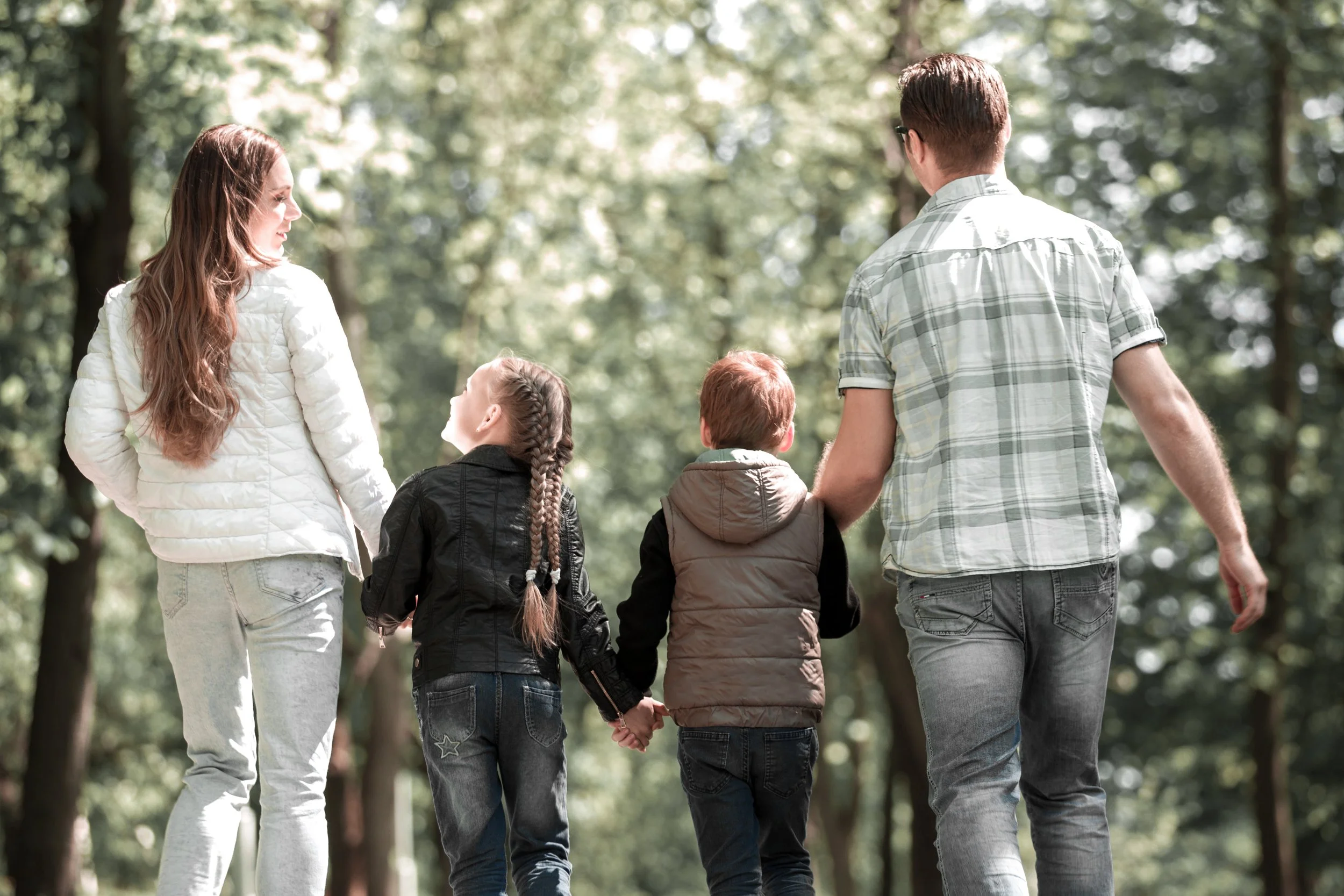 A mother and father walk hand-in-hand with their two young children in a forested park.