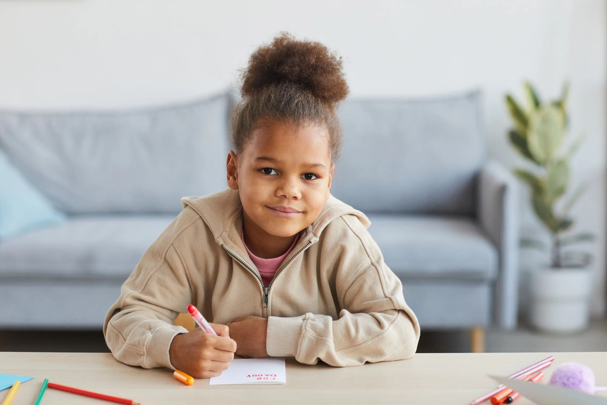 A young girl with curly hair in a bun, wearing a beige hoodie, sitting at a table with colorful markers and a card, smiling at the camera in a therapy office.