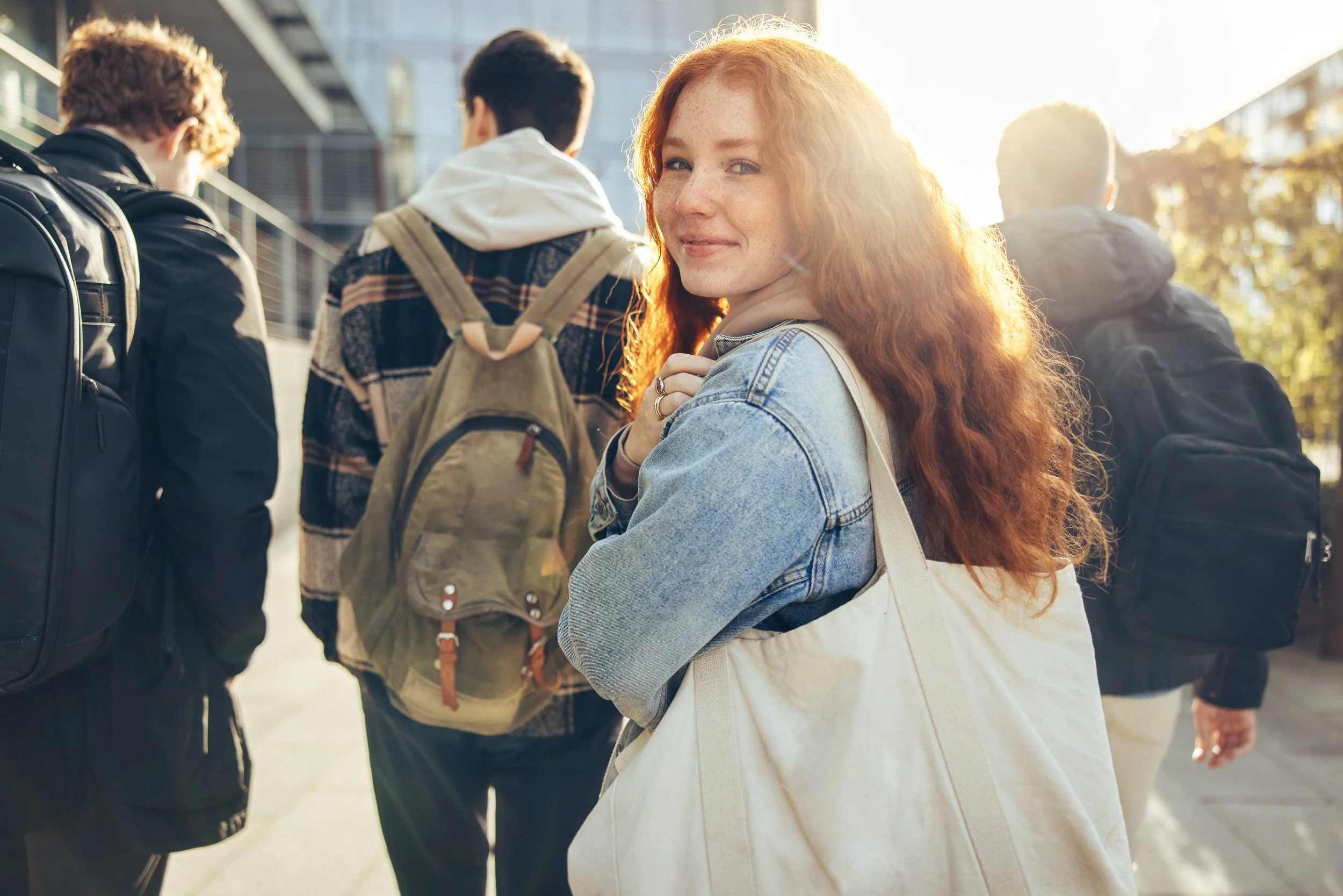 Young woman with red hair smiling and turning back to camera as she walks with three college male friends on a sunny day.