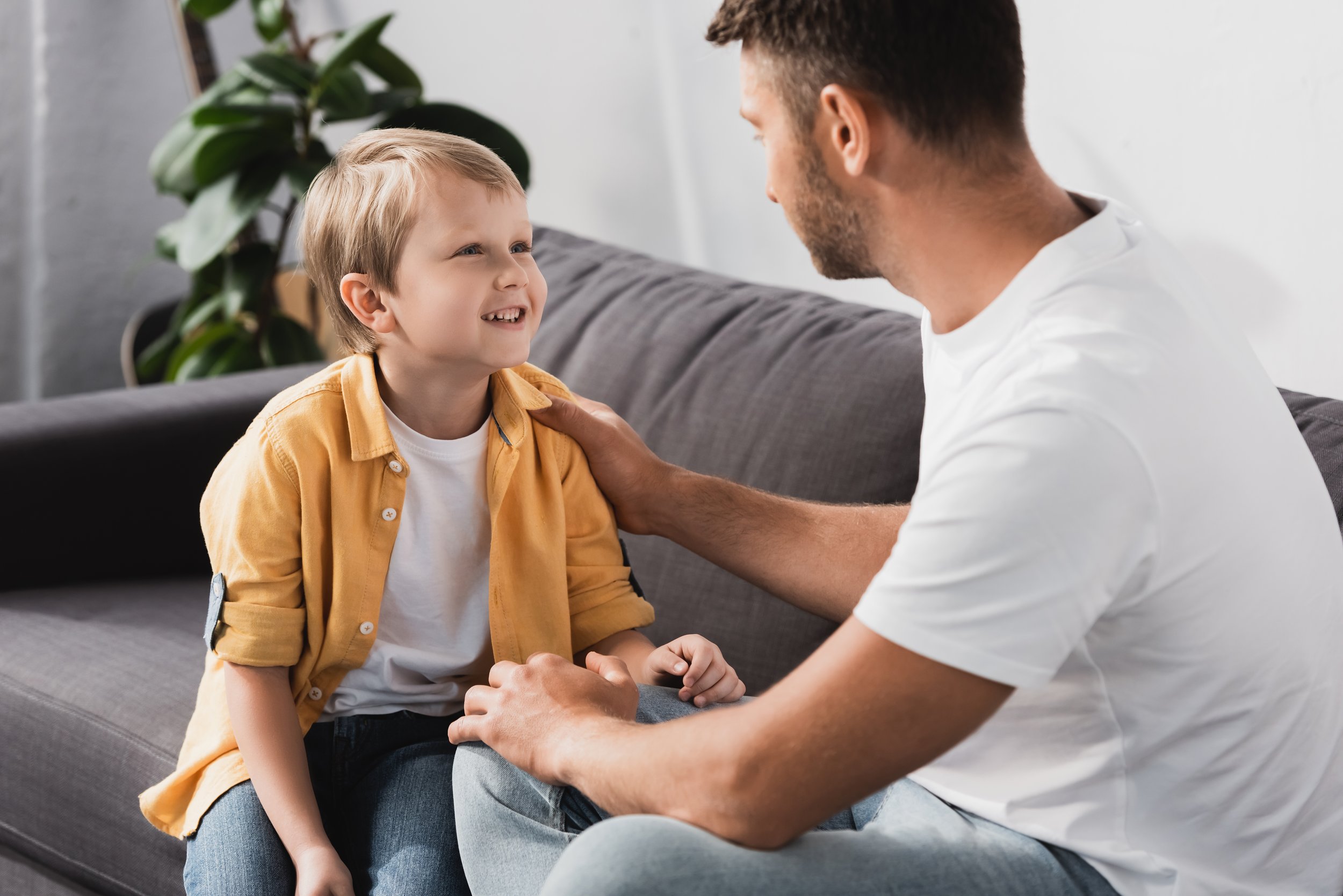 A young, smiling boy and his father sit on a couch looking at each other. The father has his right hand on his son's shoulder.