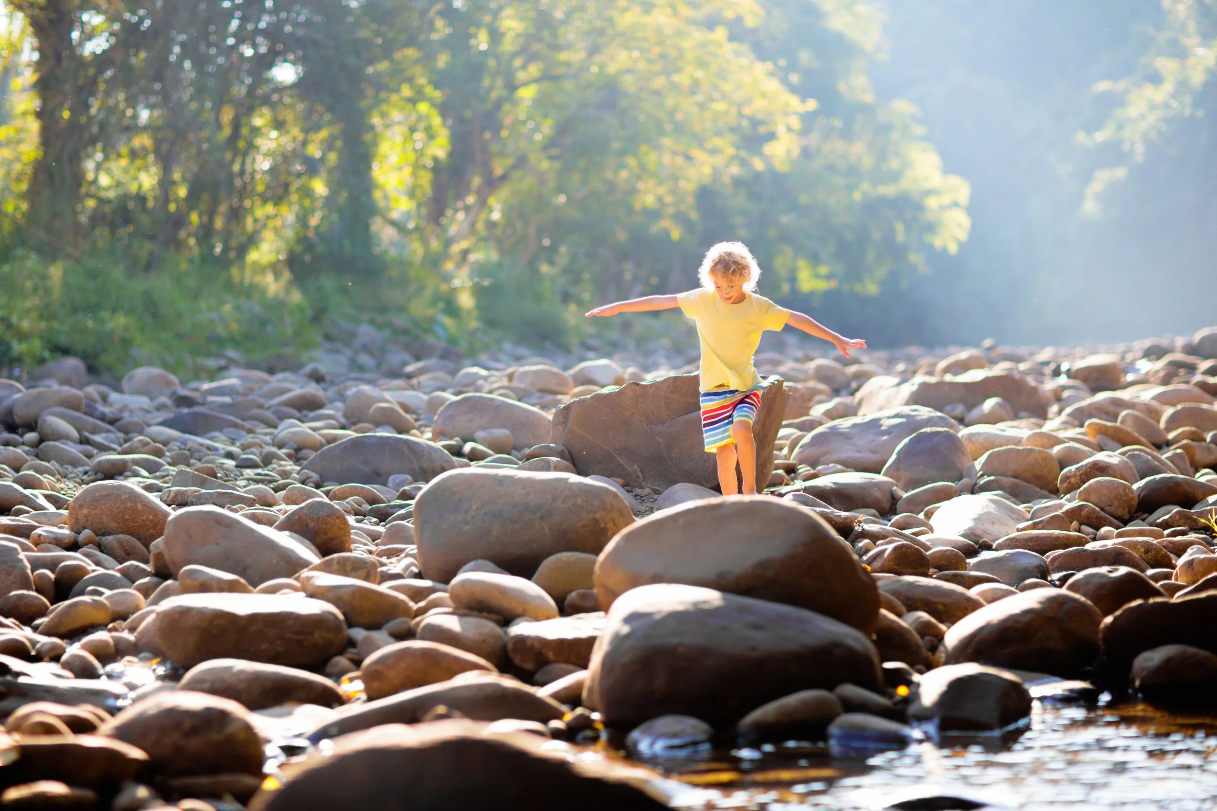 Boy in yellow shirt and striped shorts balancing on a large rock among many stones by a river with sunlight filtering through trees in the background.