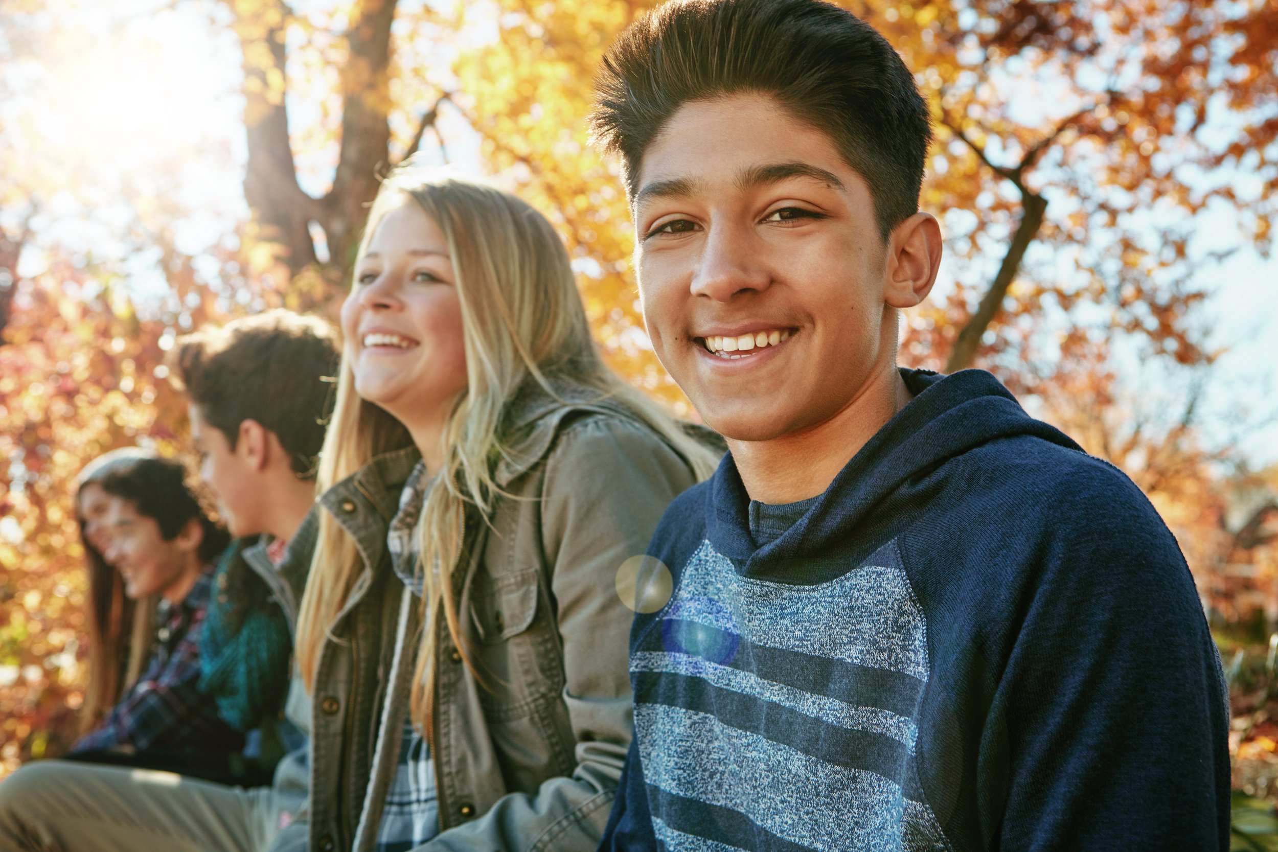Smiling teenagers sitting in a park under trees with golden, fall color leaves.