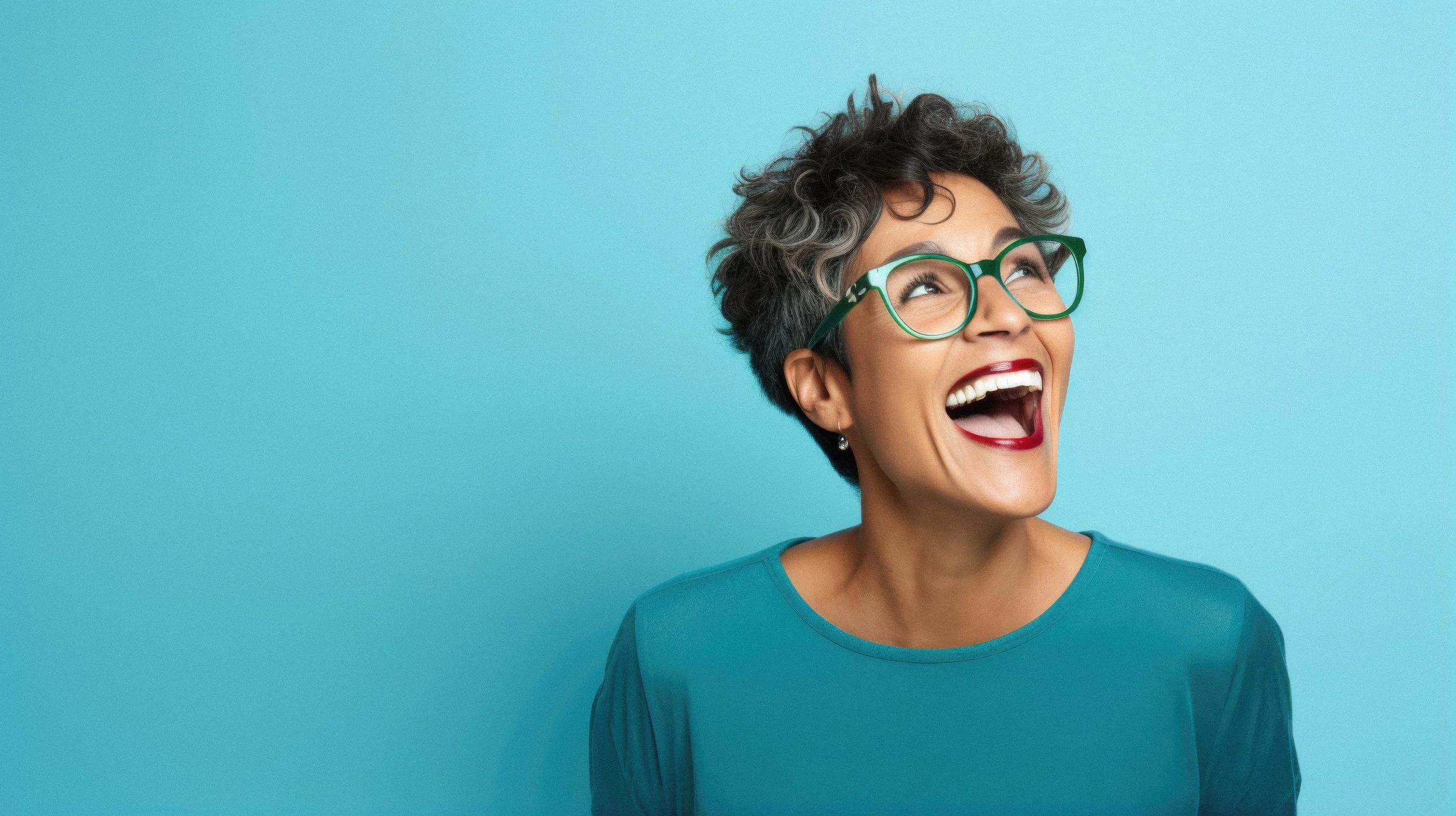 A woman with short gray curly hair, wearing green glasses and a teal shirt, is laughing joyfully against a light blue background.