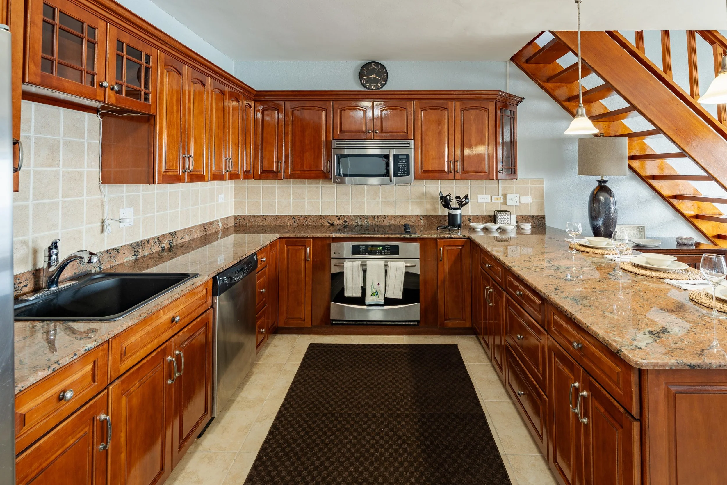Kitchen with wooden cabinets, granite countertops, and stainless steel appliances, including a stove, microwave, and dishwasher. There is a black sink, a clock on the wall, and a staircase with wooden steps ascending to the right. A dining table with