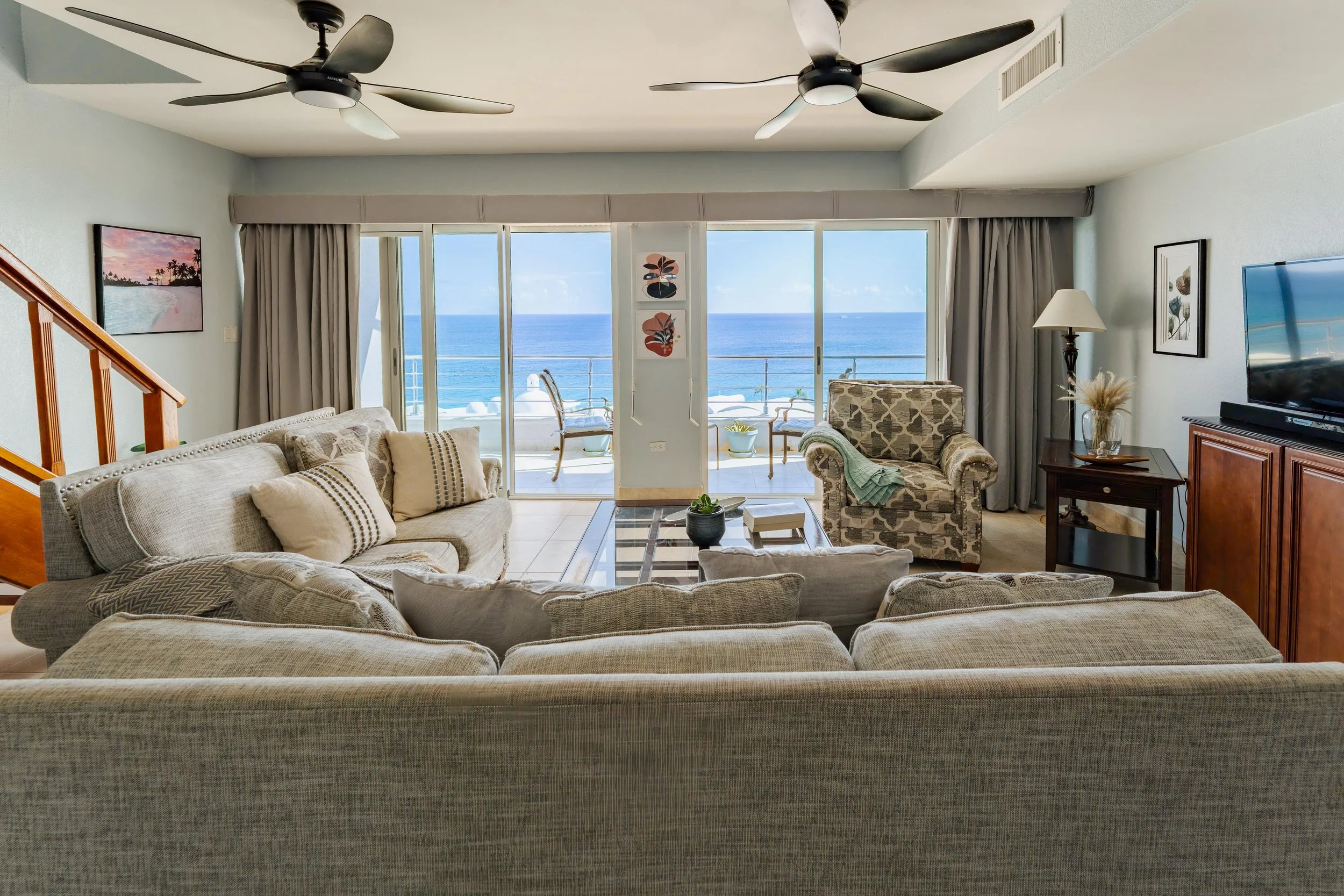 Living room with beige sofas, patterned armchair, coffee table, TV on wooden cabinet, sliding glass doors leading to a balcony with ocean view, ceiling fans, artwork, and curtains.