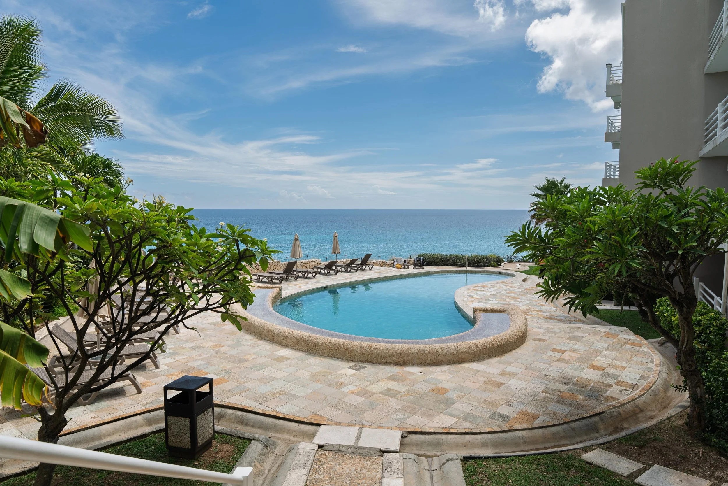 Swimming pool area overlooking the ocean with lounge chairs and umbrellas, surrounded by tropical plants and buildings.