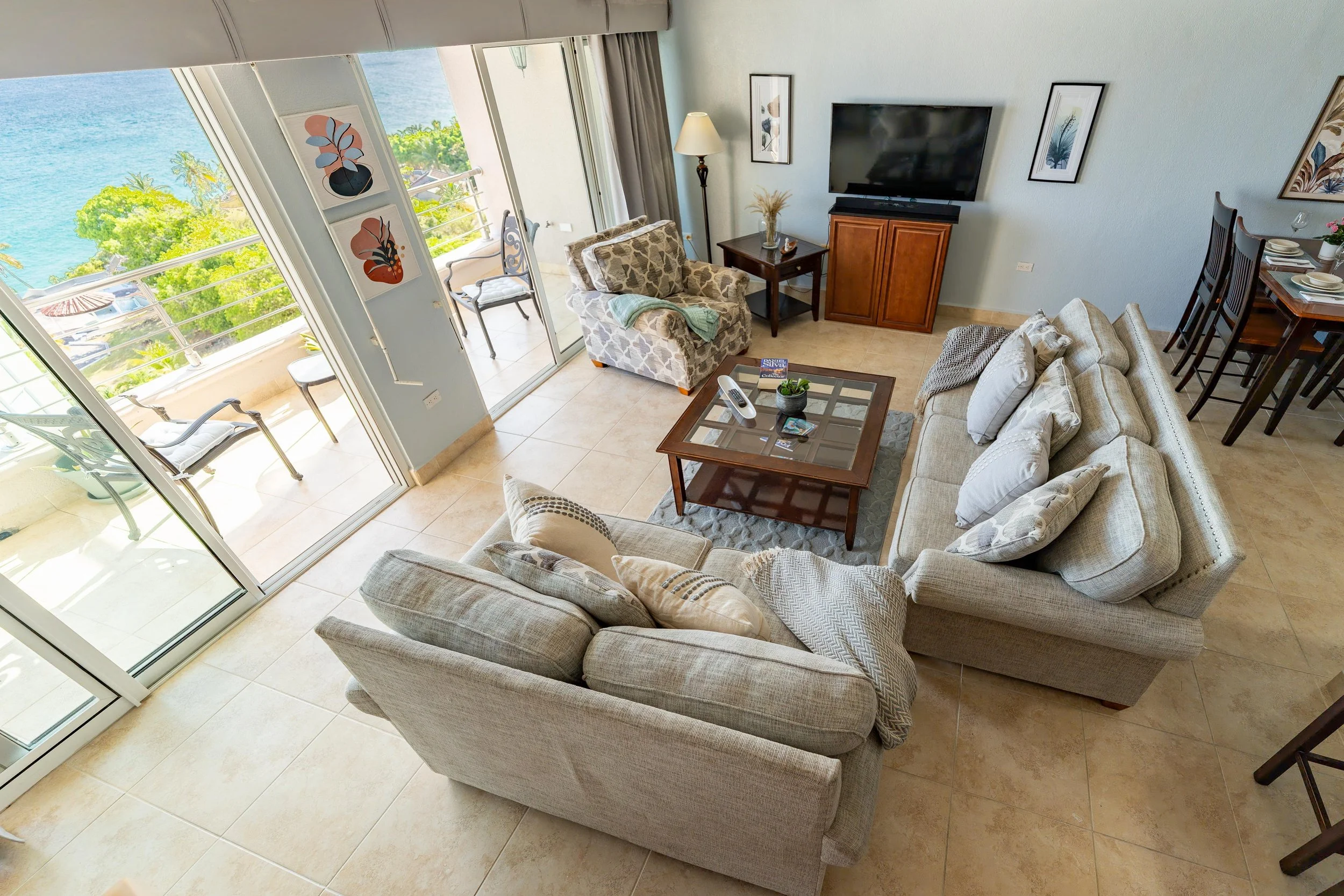 Living room with beige sofas and armchair, glass-topped coffee table, and ocean view balcony with chairs.
