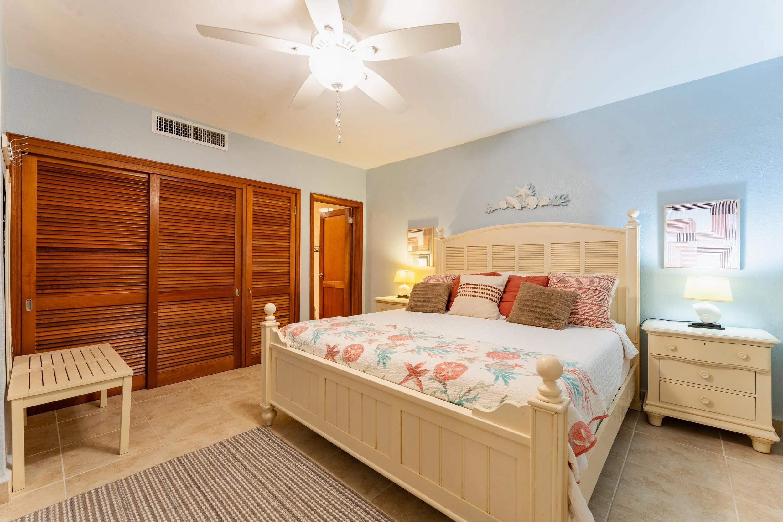 Cozy bedroom with a white wooden bed, tropical decor bedding, and multiple pillows, two nightstands with lamps, wooden closet doors, and a ceiling fan with light.