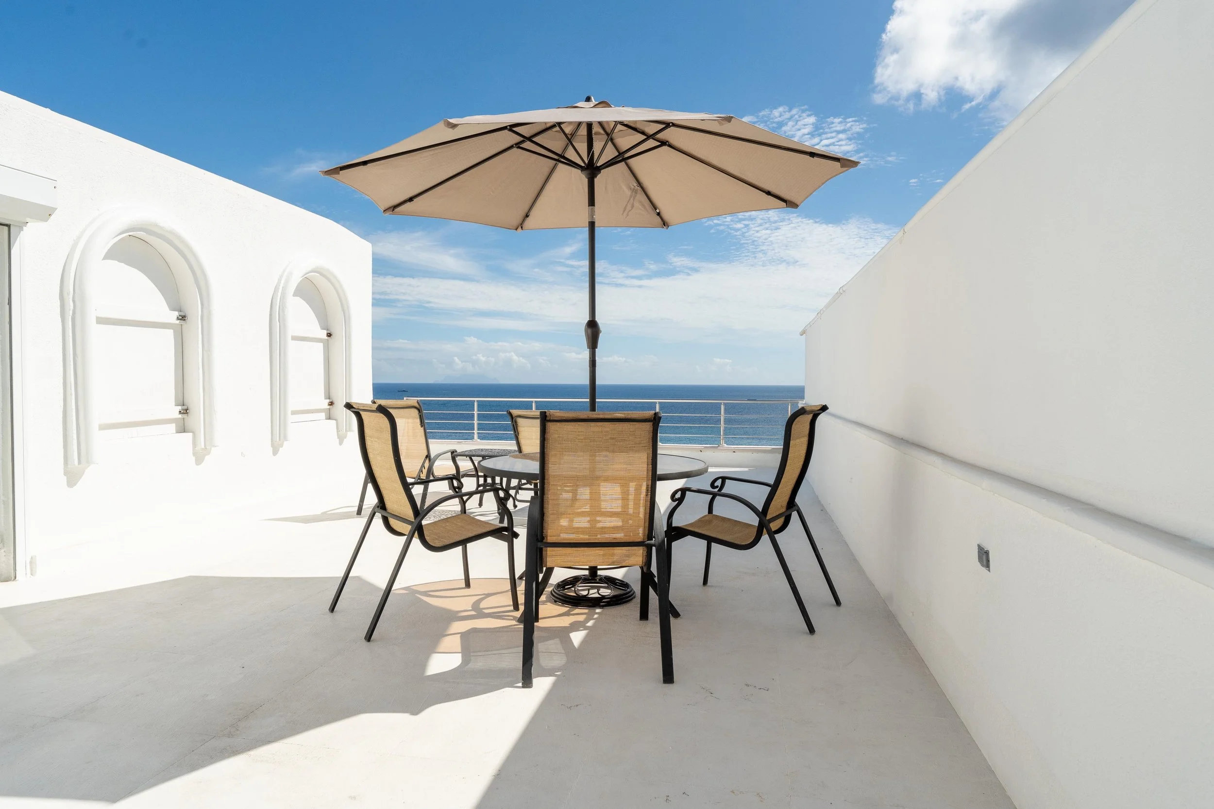 Outdoor patio with a round table, six chairs, a large beige umbrella, white walls, and an ocean view in the background.