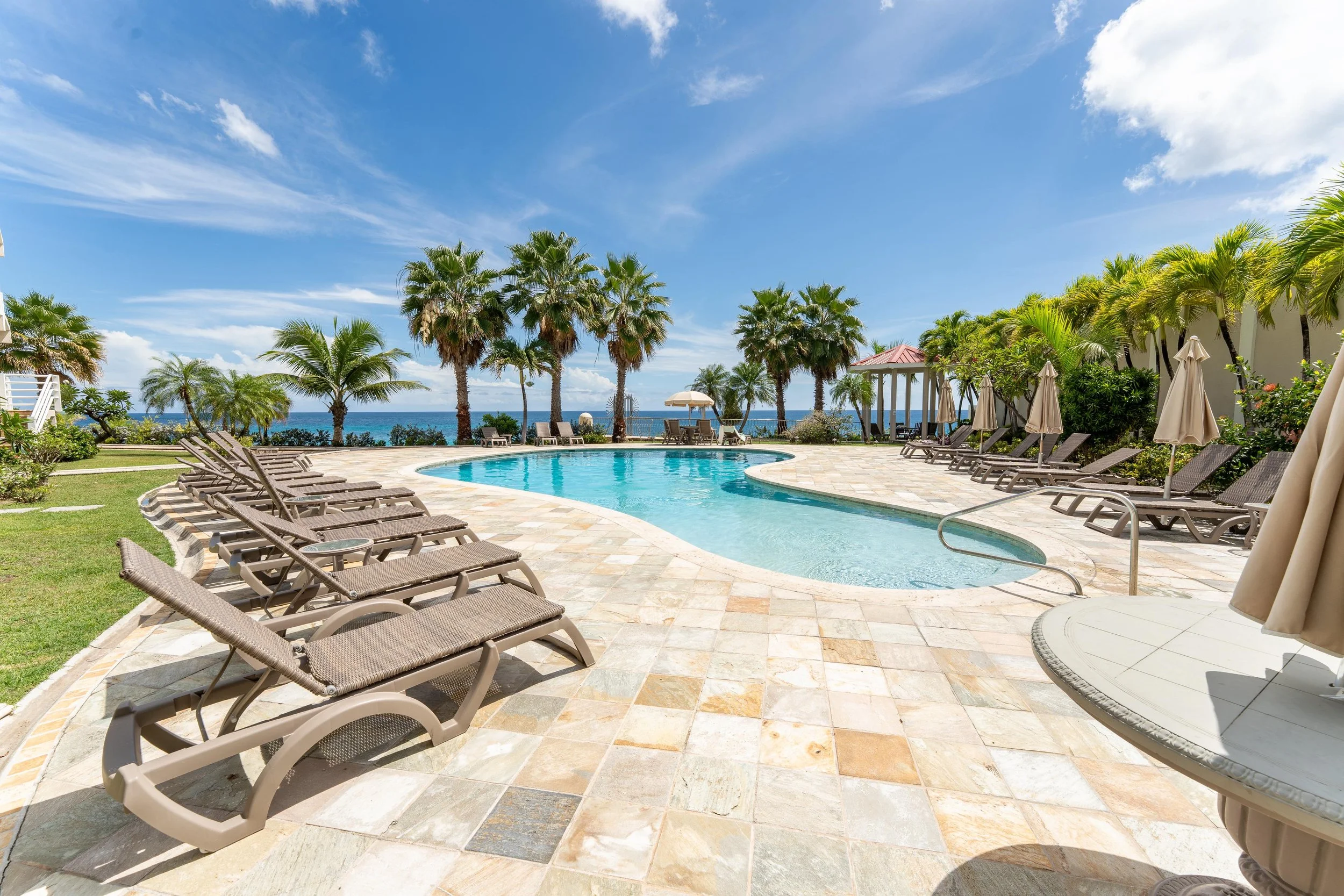 Swimming pool with lounge chairs and umbrellas, palm trees, and ocean view on a sunny day.