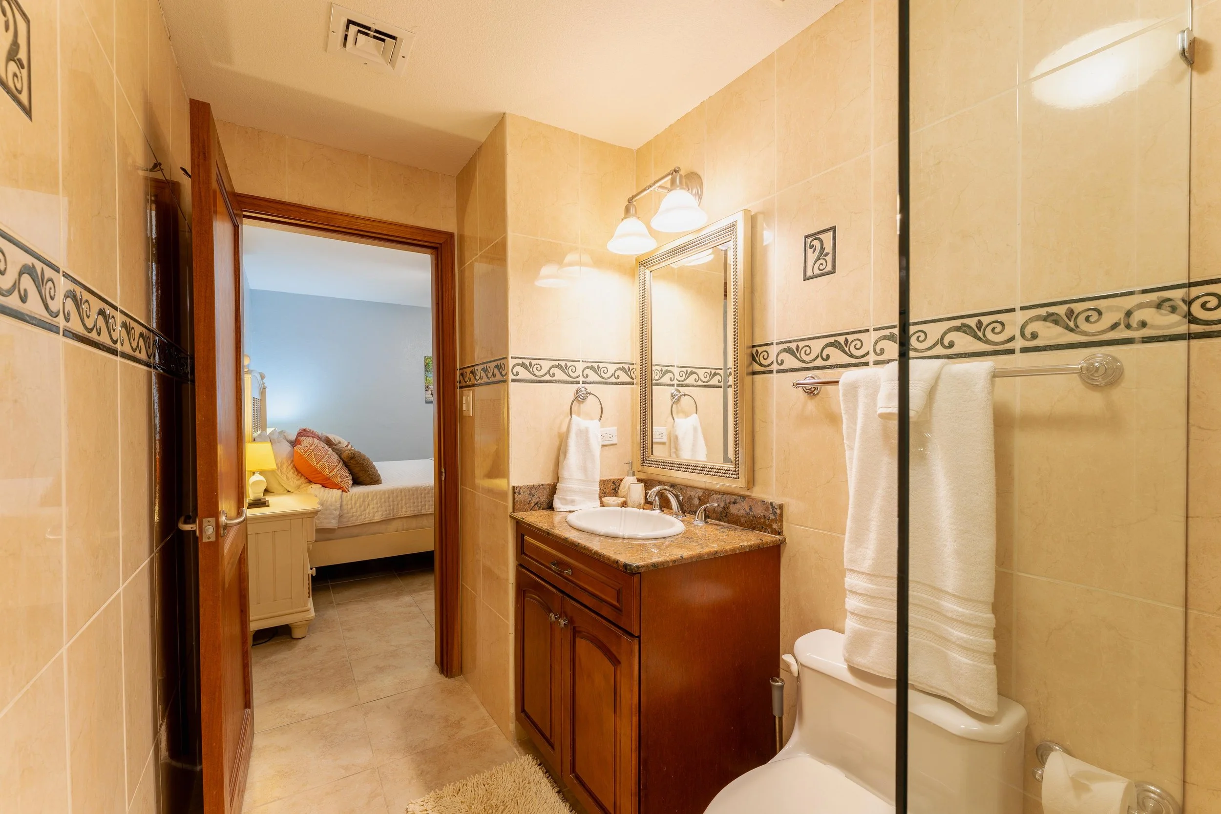 Bathroom with beige tiles, a wooden vanity with a granite countertop, a mirror, towels, a toilet with a towel on top, and a partial view of a bedroom through the open door.