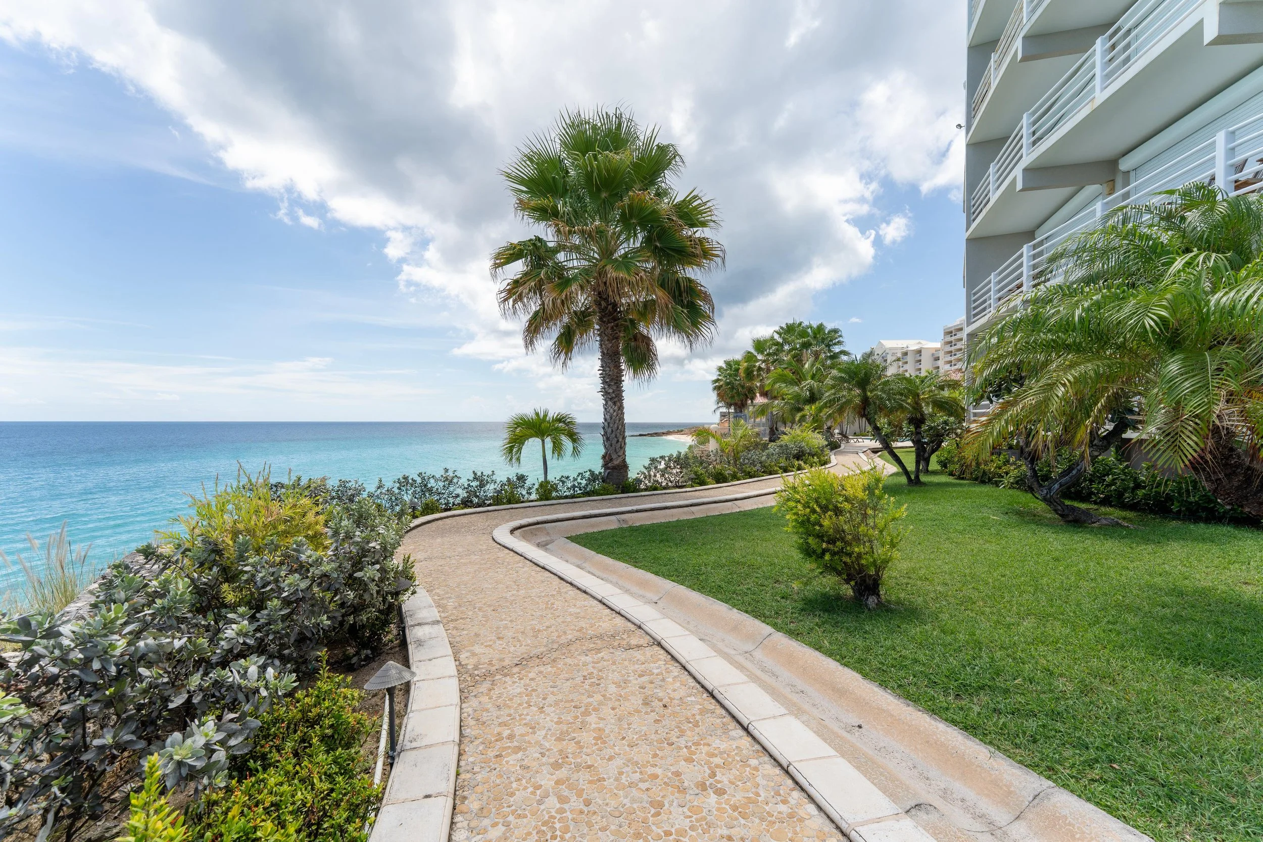 A curved pathway along a tropical garden with palm trees, overlooking the ocean, with a multi-story white building to the right, partly cloudy sky.