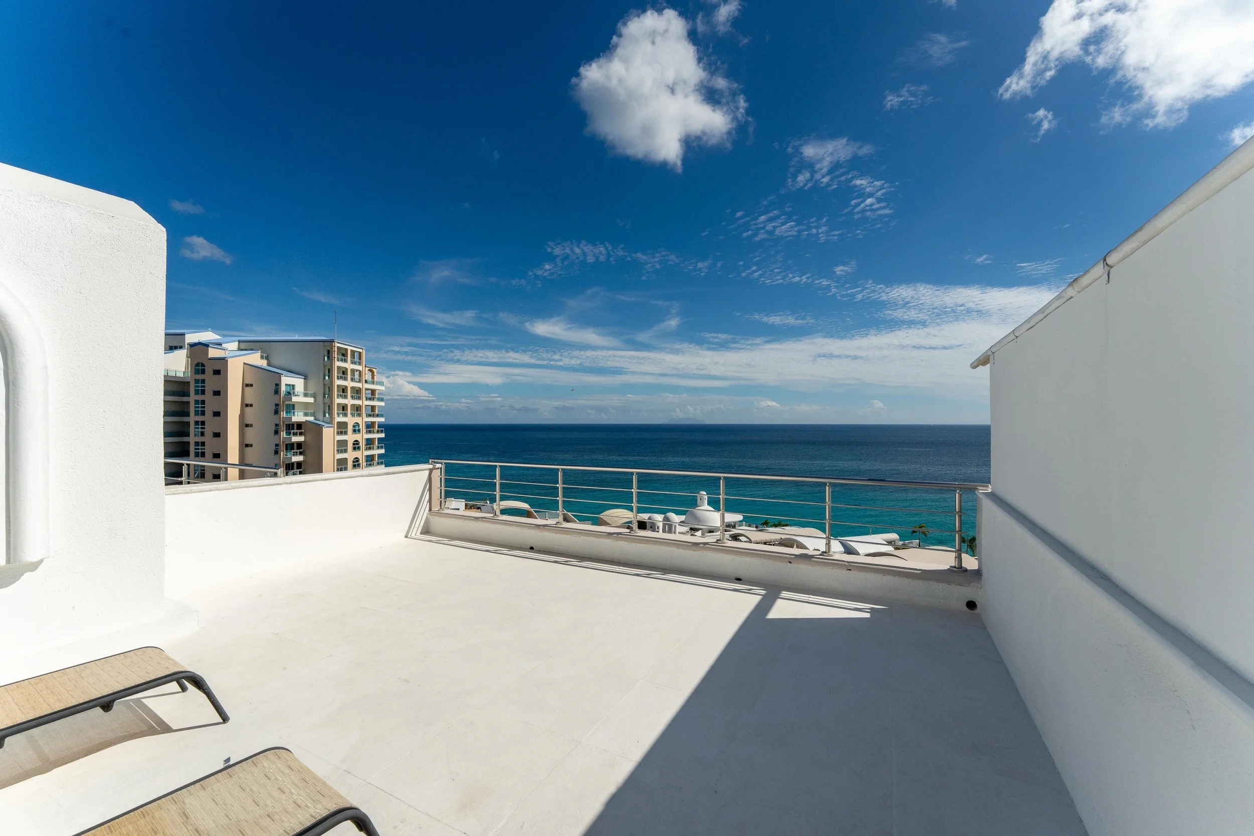 View of a balcony overlooking the ocean with white walls, a railing, lounge chairs, and a distant building under a partly cloudy sky.