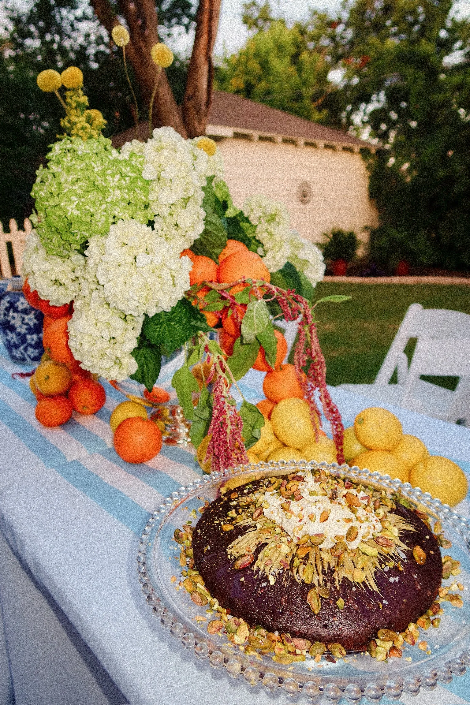 Dessert with chopped pistachios on top and a dollop of whipped cream on a glass cake stand, alongside a floral centerpiece of white hydrangeas and orange and yellow fruits on a table with a blue and white striped tablecloth.