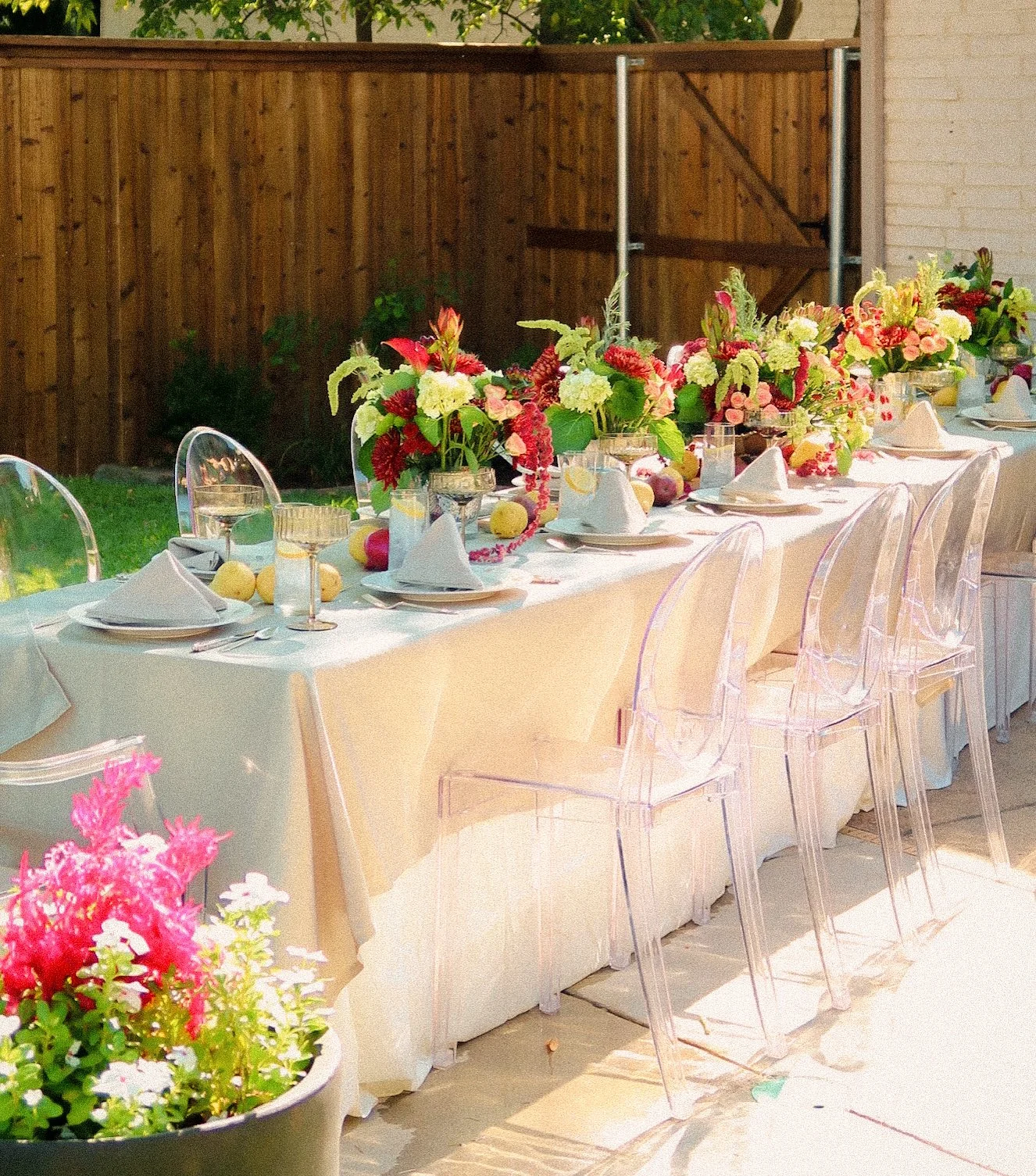An outdoor dining table decorated with floral centerpieces and set for a meal, with clear acrylic chairs, white napkins, and glassware, with a wooden fence and greenery in the background.