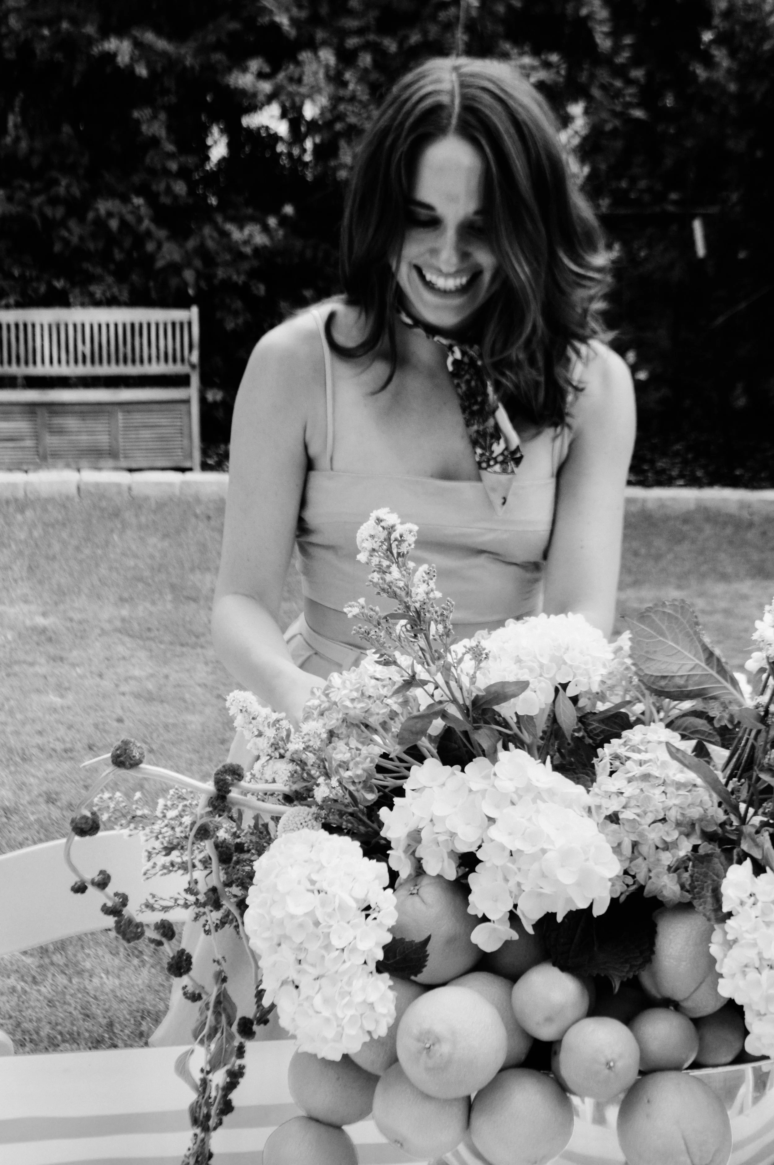 A woman smiling and looking down at a large floral arrangement of hydrangeas, apples, and other flowers, outdoors with a bench and trees in the background.
