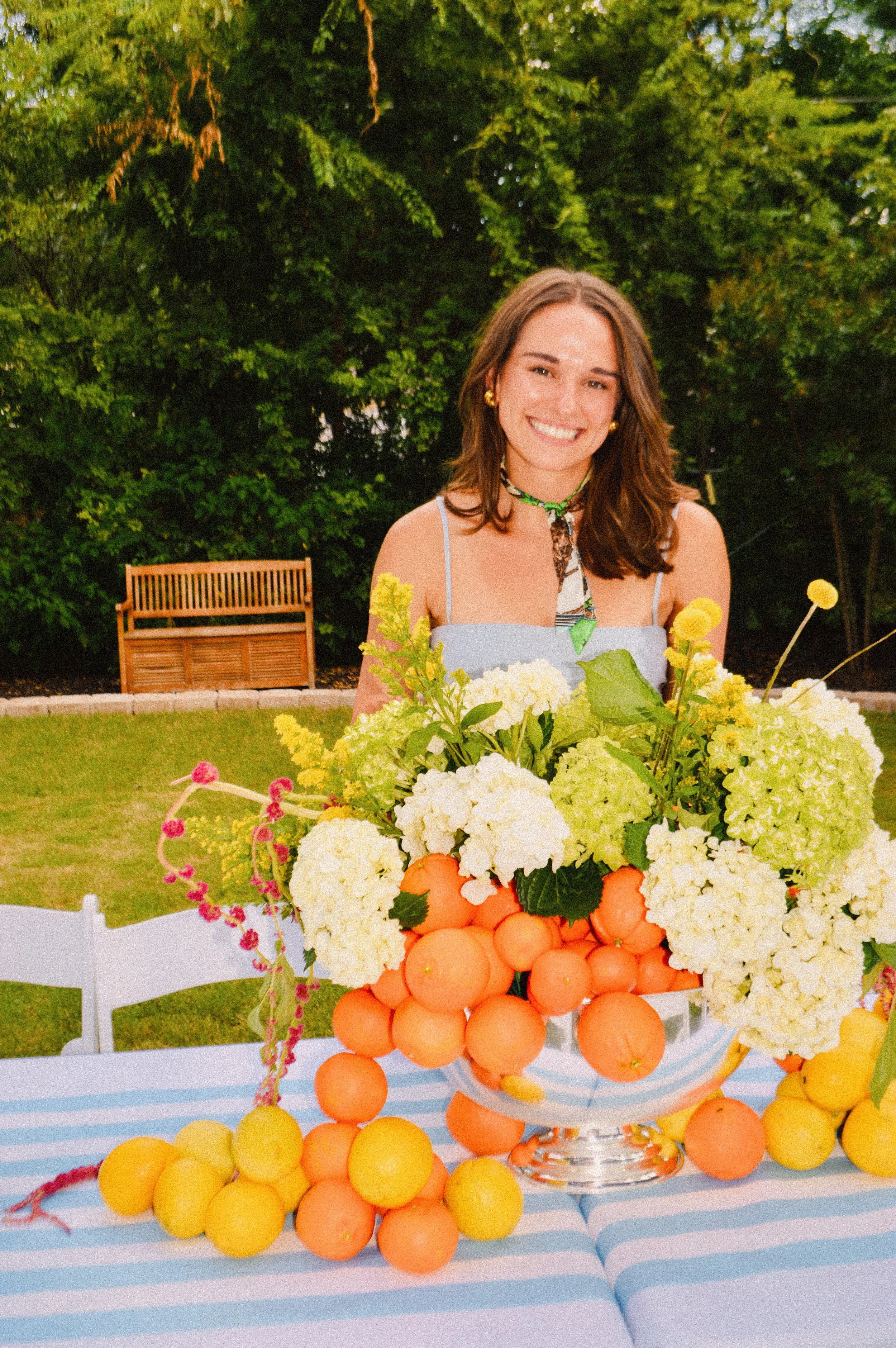 A smiling woman with brown hair is standing behind a table decorated with a floral arrangement of white hydrangeas, green leaves, and yellow and pink flowers, surrounded by oranges and lemons, outdoors with greenery and a bench in the background.