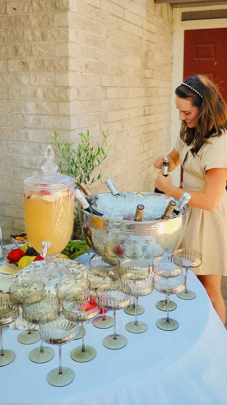 A woman in a beige dress and pearl headband pouring drinks into glasses at a celebration with a table of champagne, fruit, and a large ice bucket filled with bottles.