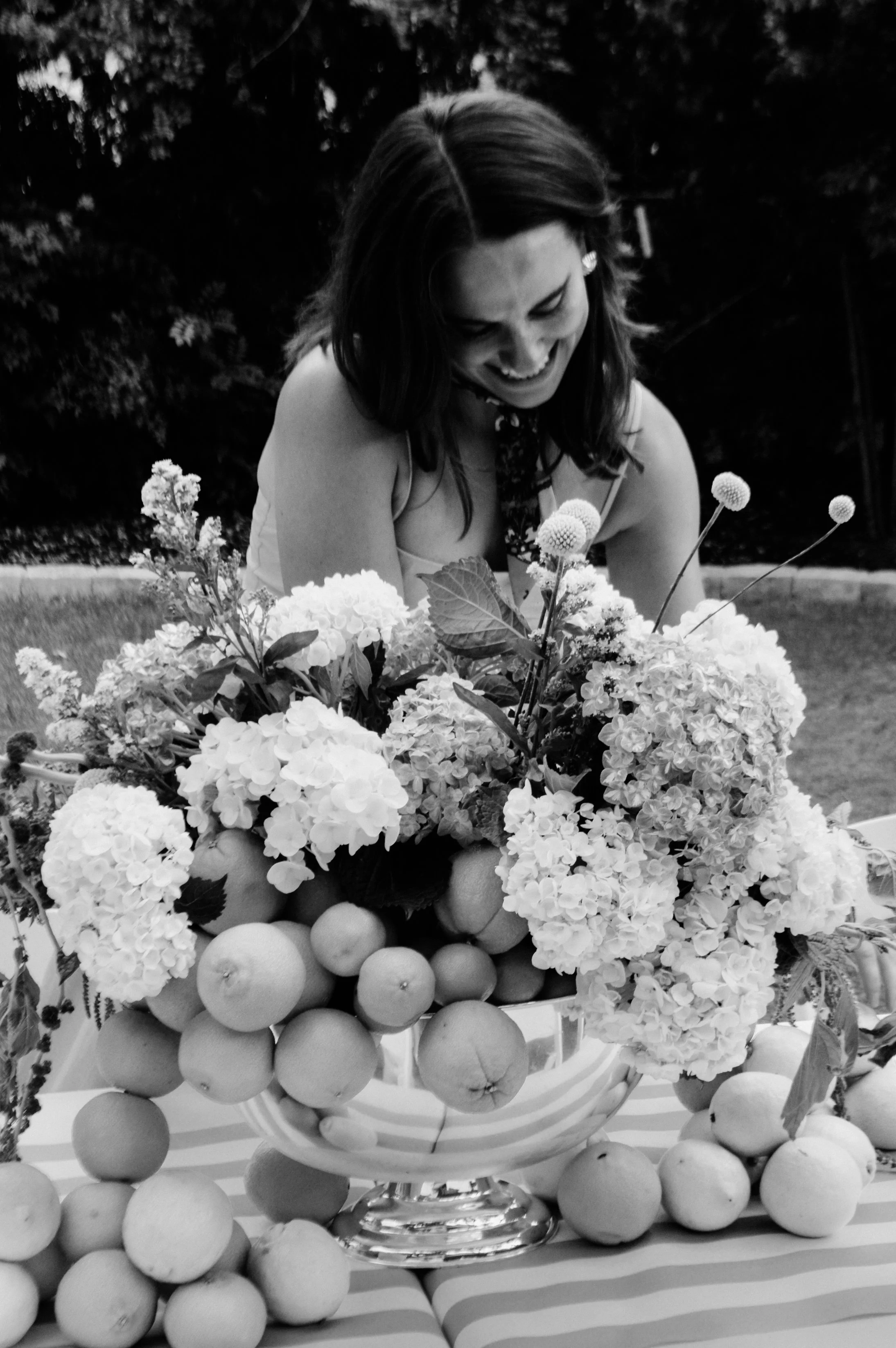 A woman arranging a floral display with hydrangeas, flowers, and apples on a table outdoors.