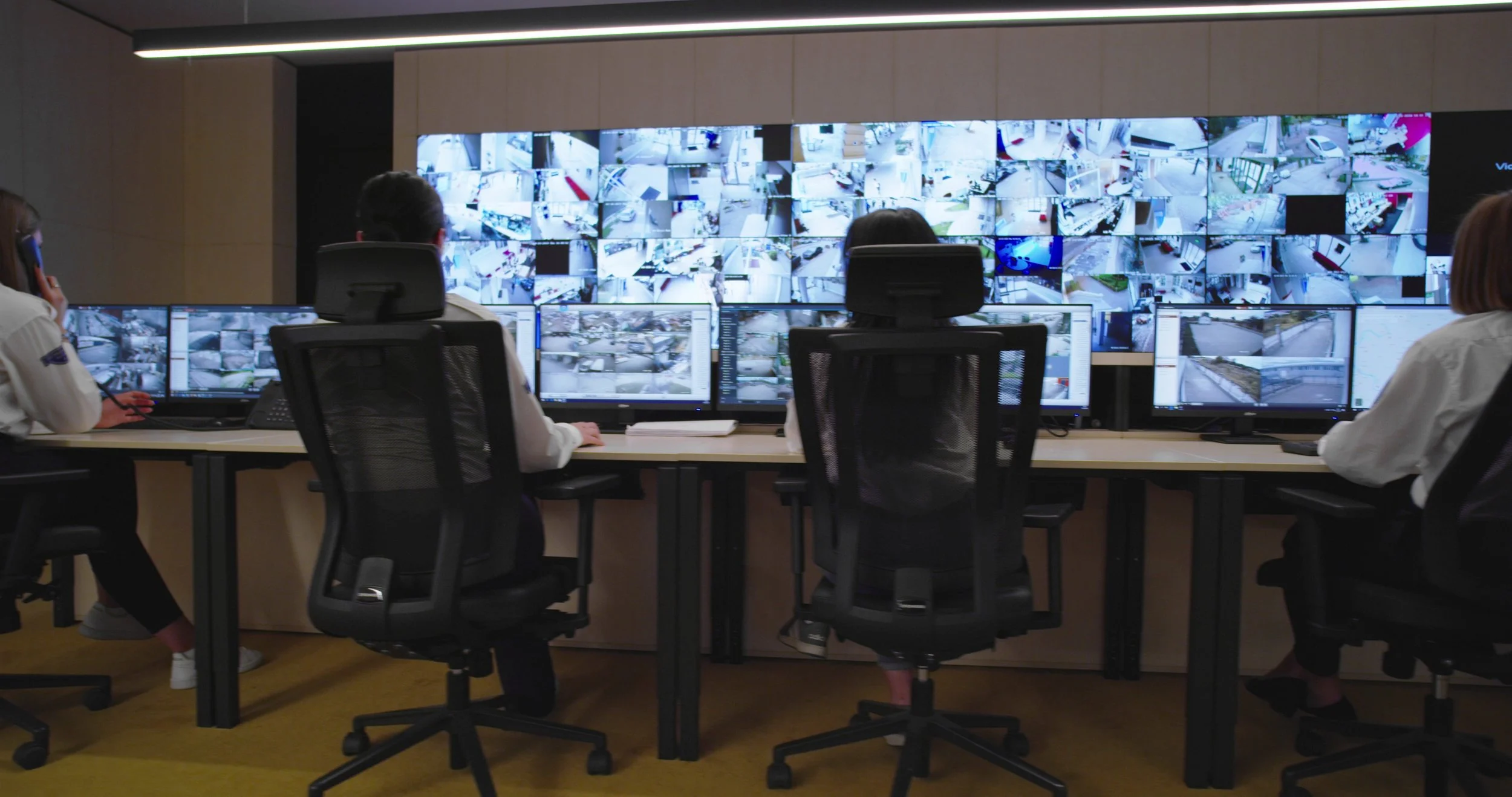 Security control room with multiple surveillance monitors, three people monitoring screens, and large video wall displaying CCTV footage.