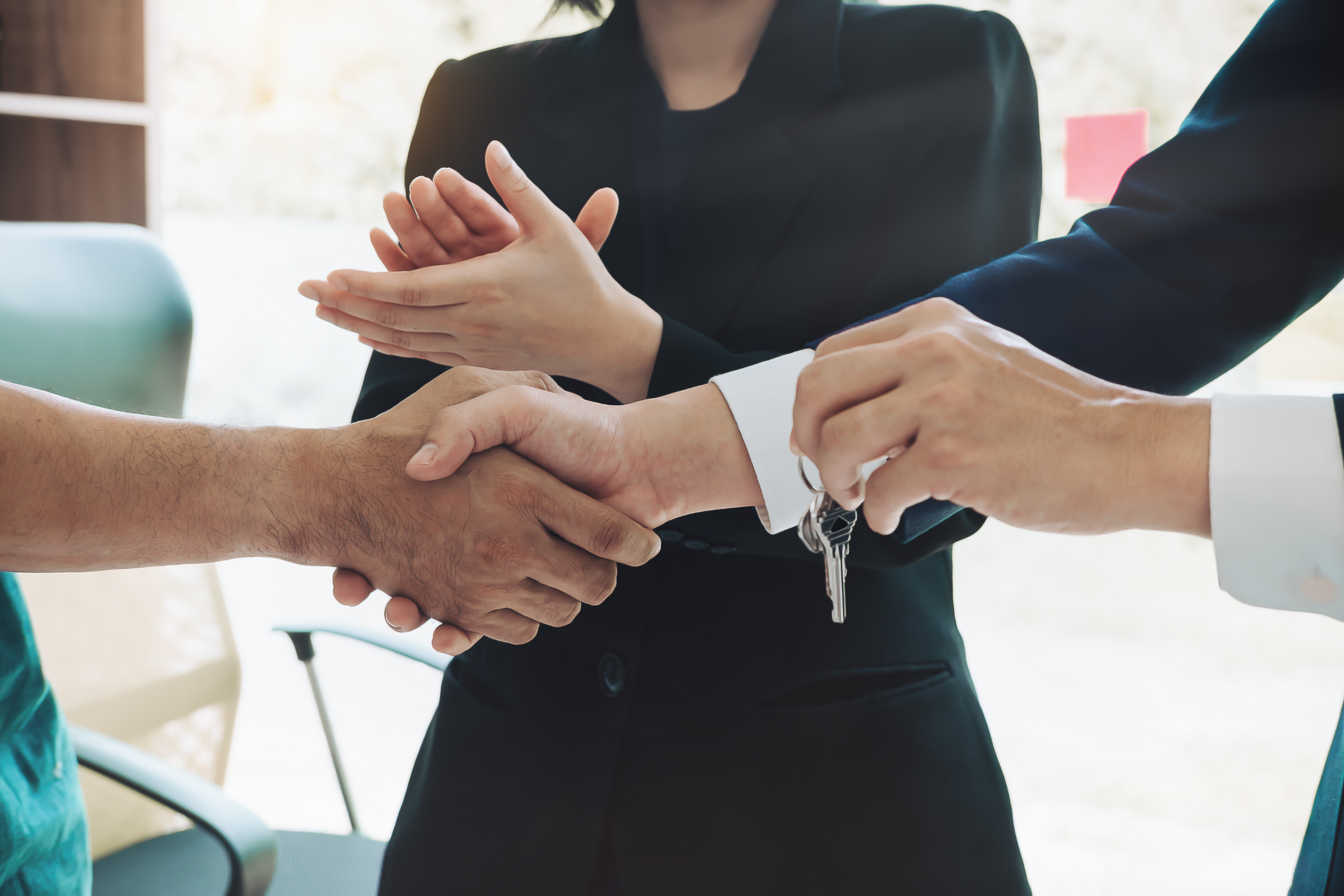 A person in a black suit is handing over a set of keys and shaking hands with another individual in a professional setting, suggesting a transaction or agreement.