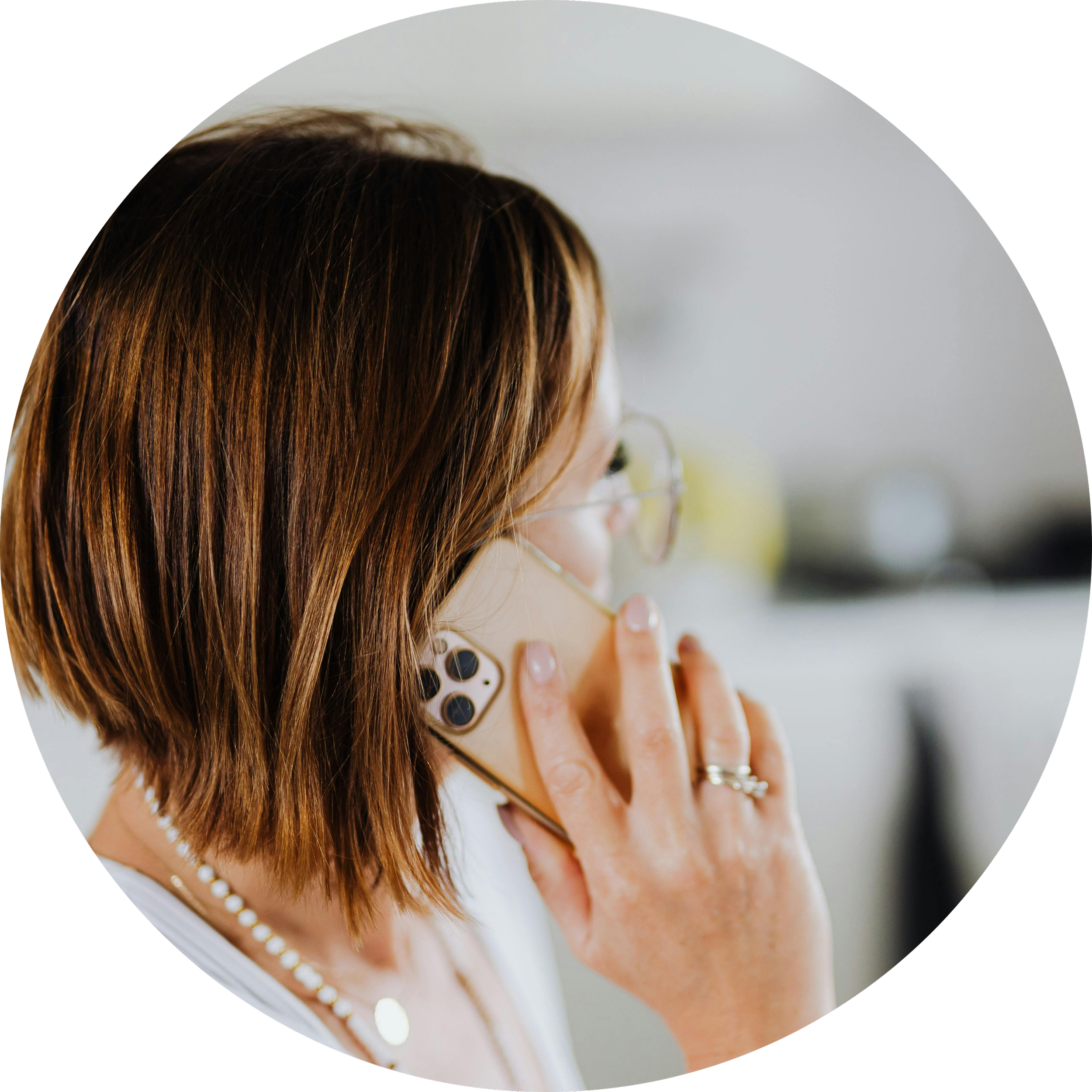 A woman with shoulder-length brown hair, glasses, and a pearl necklace, talking on her smartphone.