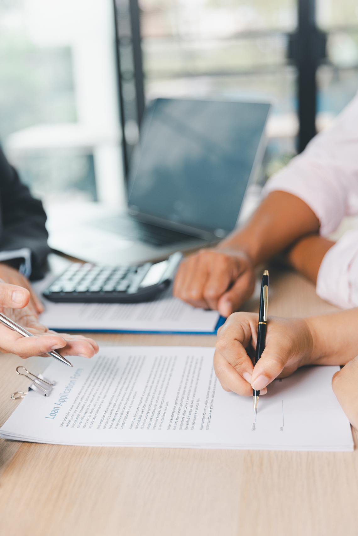 Two people shaking hands over documents, with a laptop and calculator on the table, in a bright office setting.