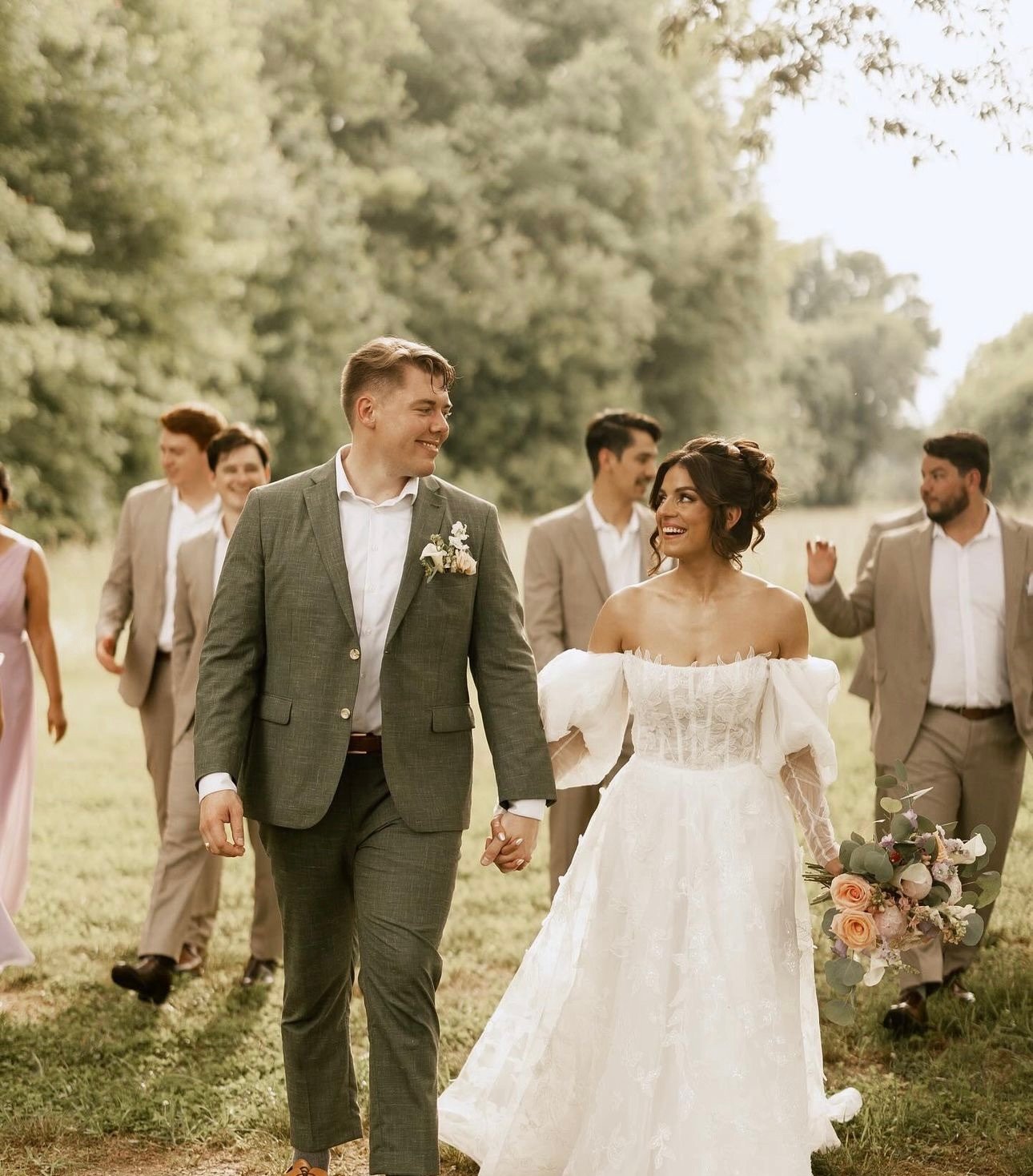 A bride and groom walking hand in hand outdoors, surrounded by friends, during a wedding celebration.