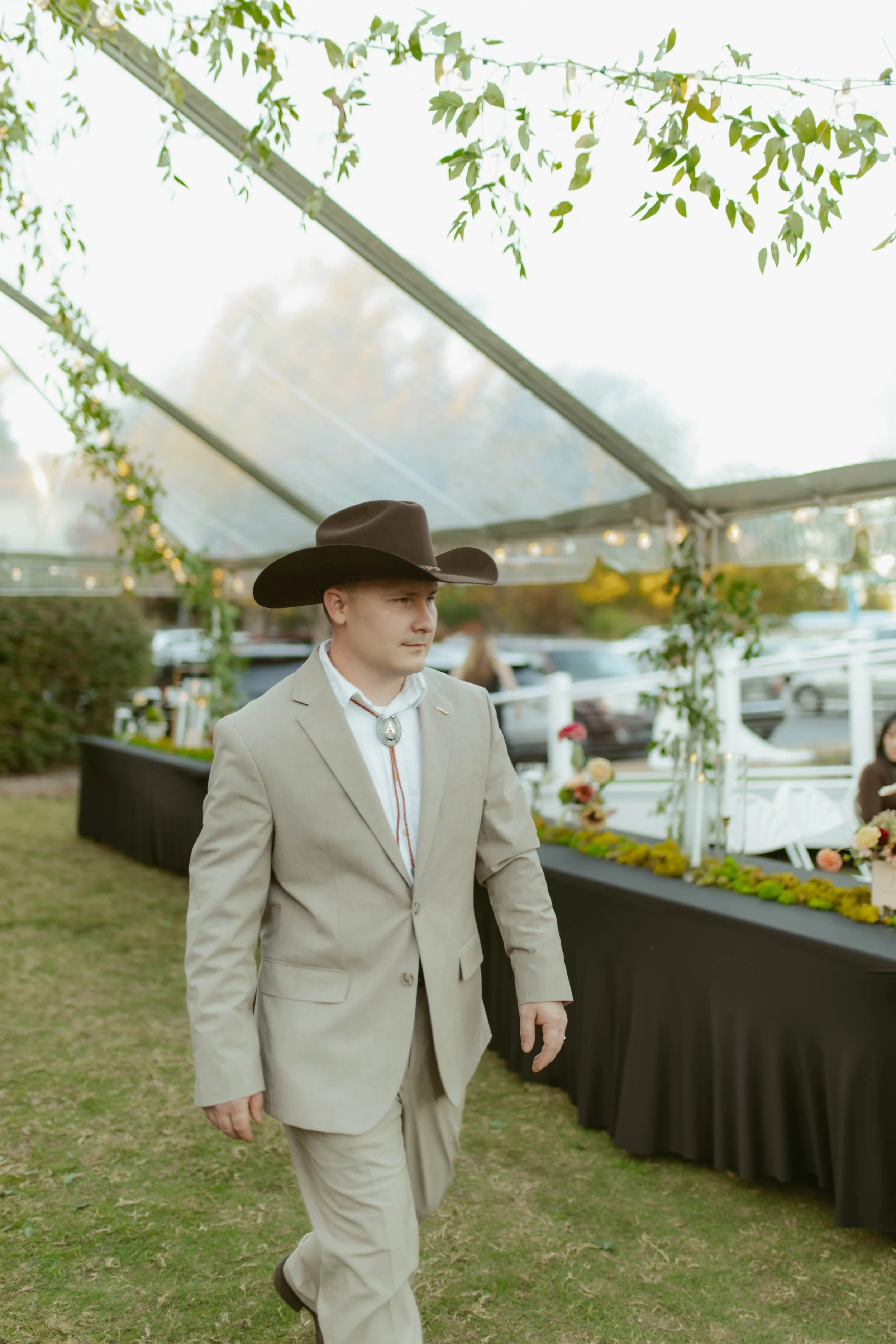 A man wearing a beige suit, white dress shirt, bolo tie, and cowboy hat walking outdoors under a clear tent decorated with greenery and string lights. There are parked cars and people sitting at tables in the background.
