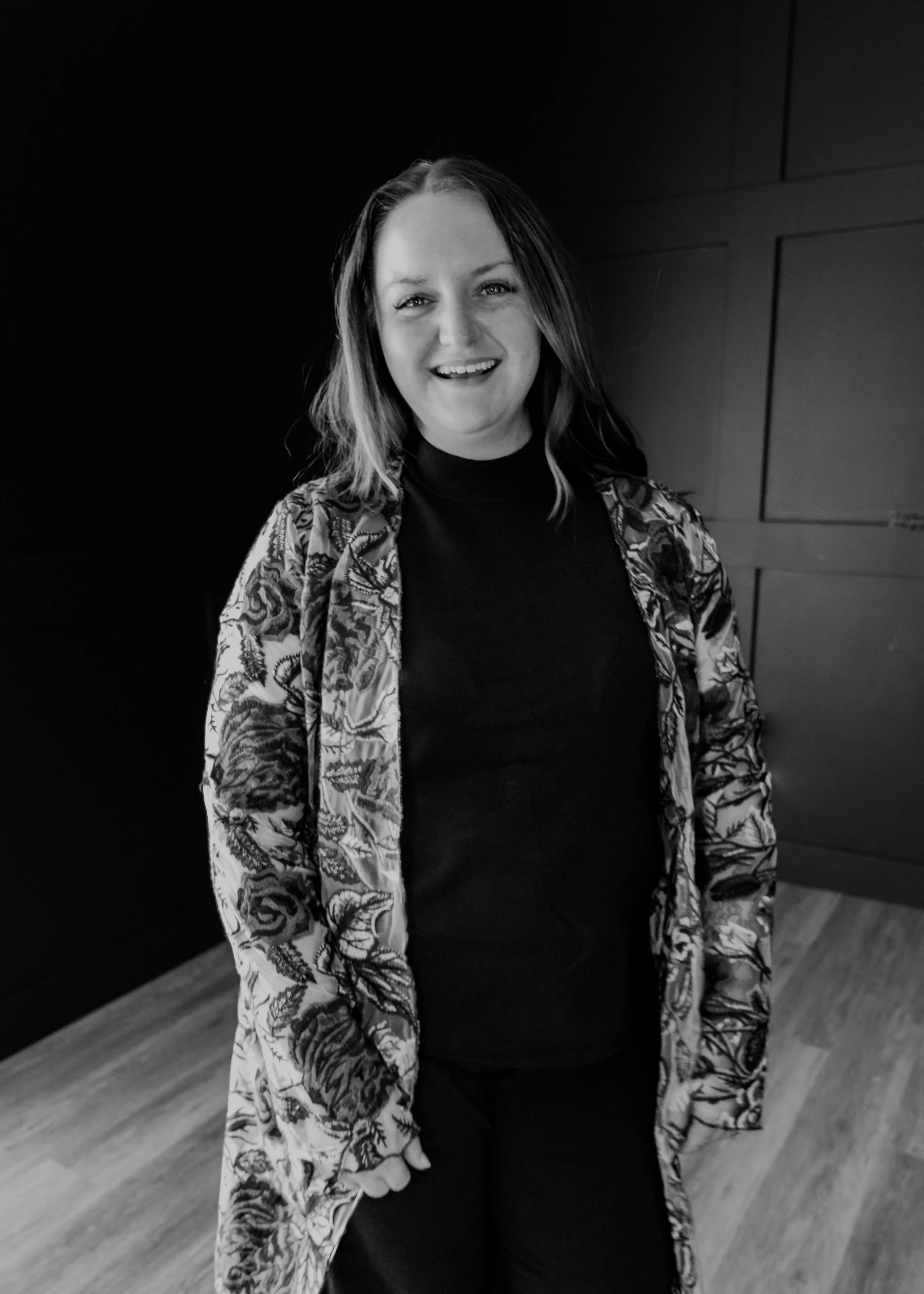 A woman with shoulder-length hair smiling, wearing a patterned jacket over a dark top, standing indoors against a dark wall.
