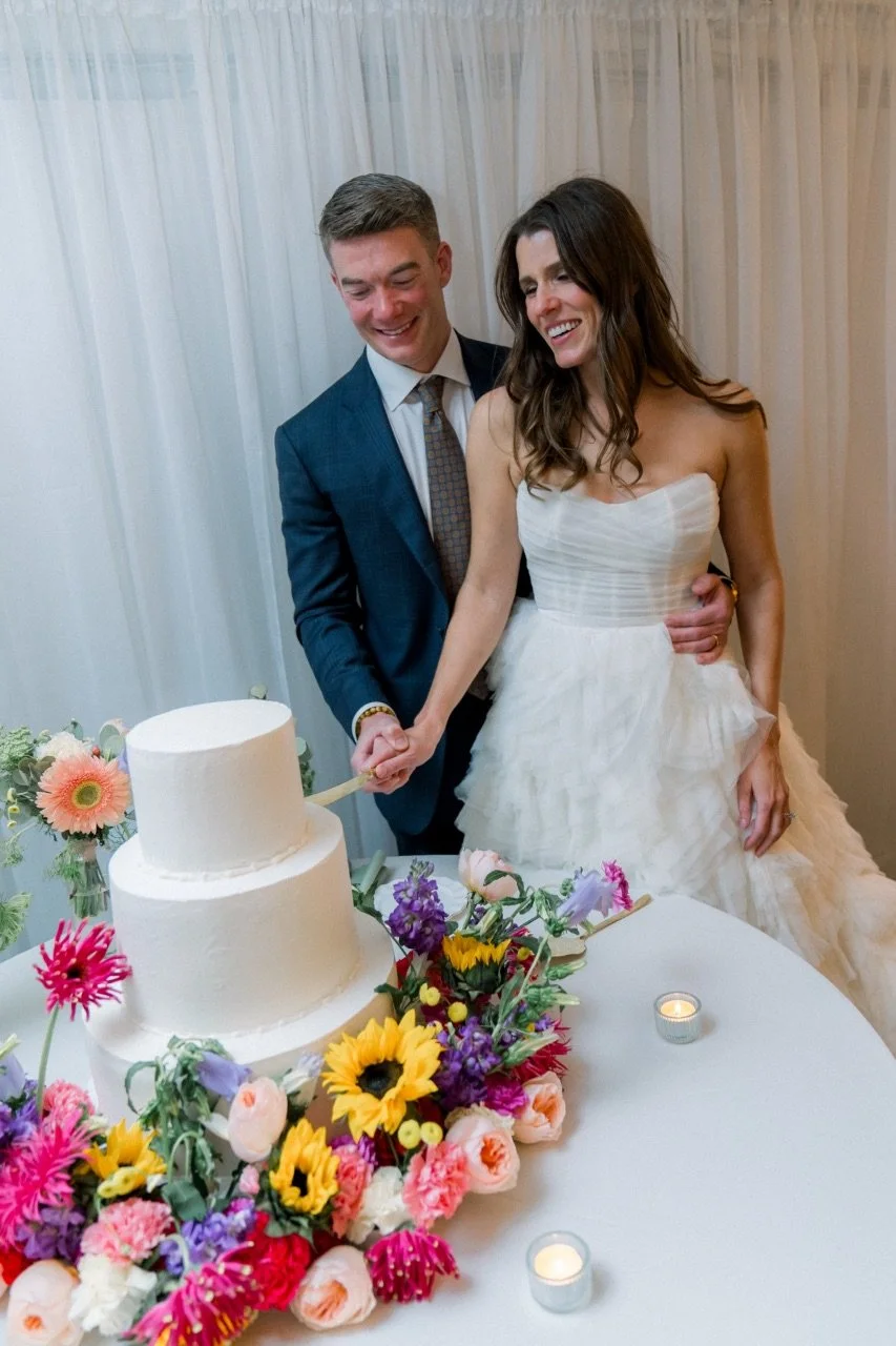 A bride and groom cut a wedding cake together, holding a knife with their hands. They are smiling and standing behind a table decorated with colorful flowers and small candles. The bride wears a strapless white wedding dress, and the groom is in a na