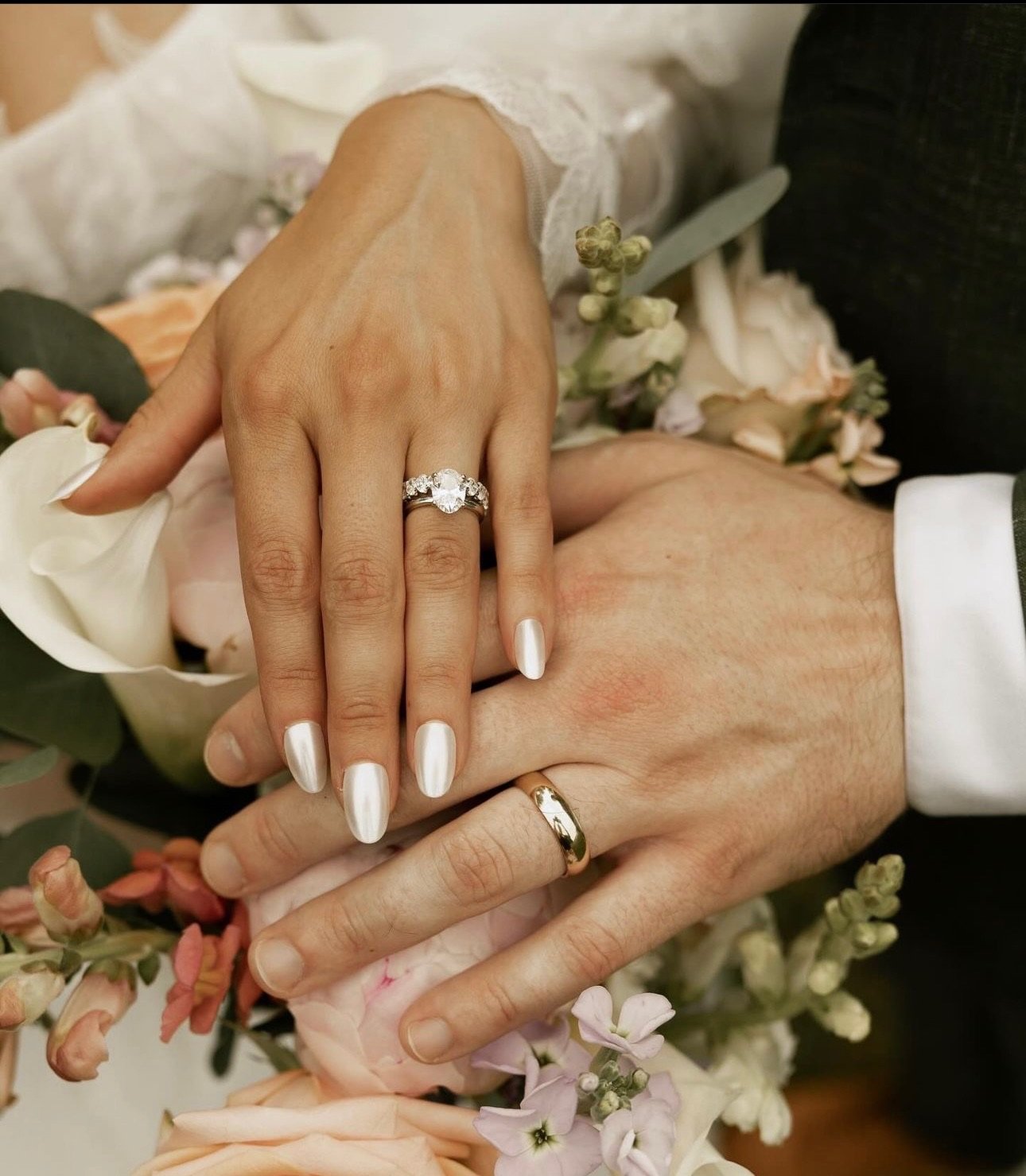 Close-up of a bride and groom's hands showing their wedding rings, with flowers in the background.