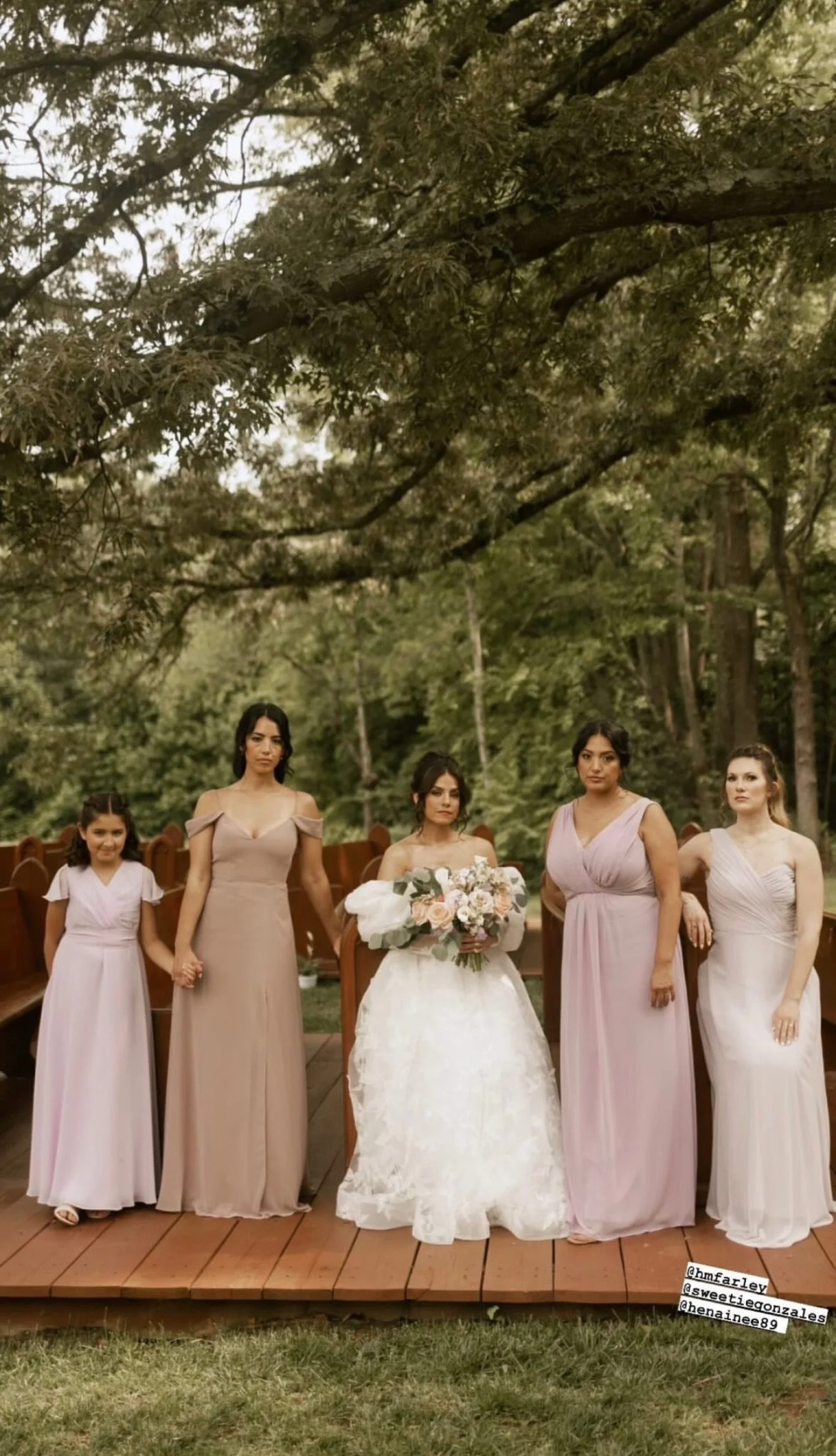 Group of five women and a girl at a wedding, standing on a wooden platform outdoors with lush green trees in the background. The bride in the center wears a white lace dress and holds a bouquet of flowers. Four women in pastel dresses and a girl in a