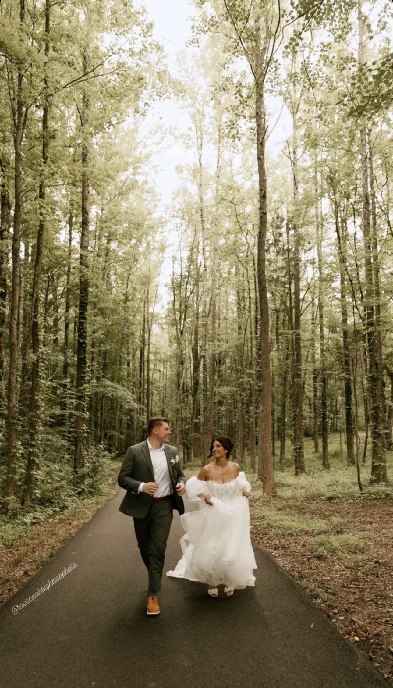 A couple in wedding attire walking through a forested park on a paved path.